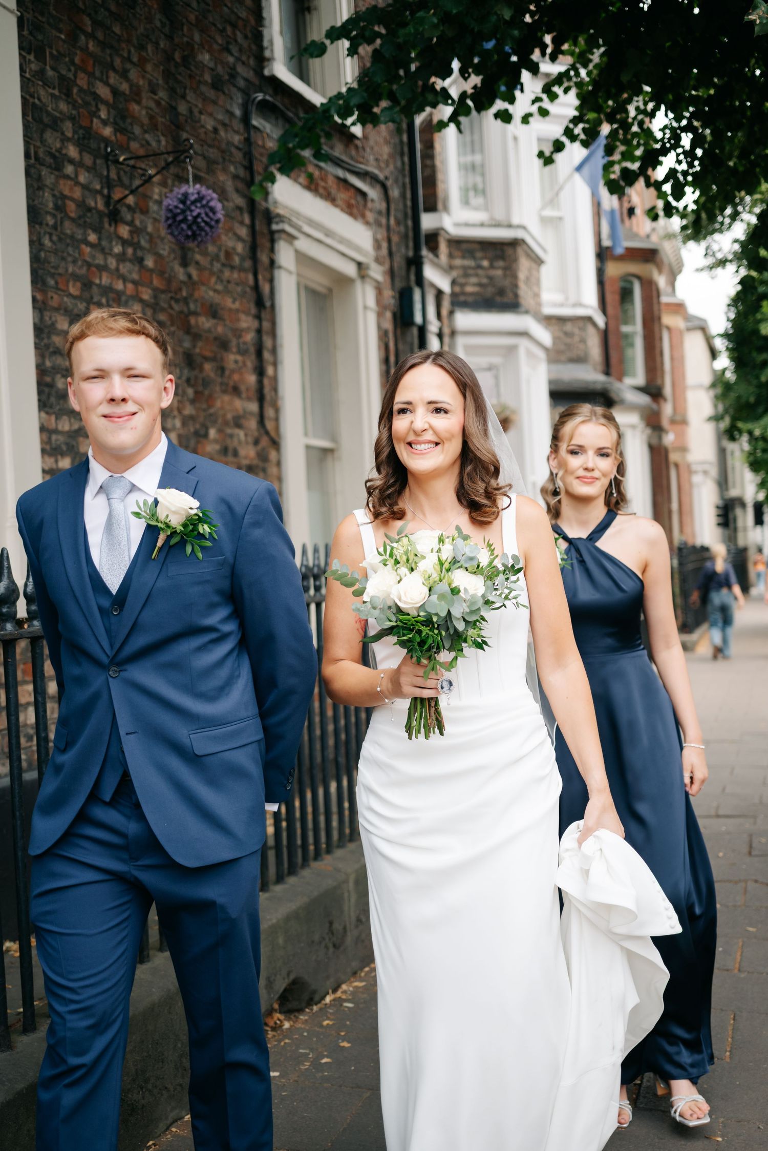 Bride in white gown holds bouquet walking with groomsman in navy suit and bridesmaid on a city street.