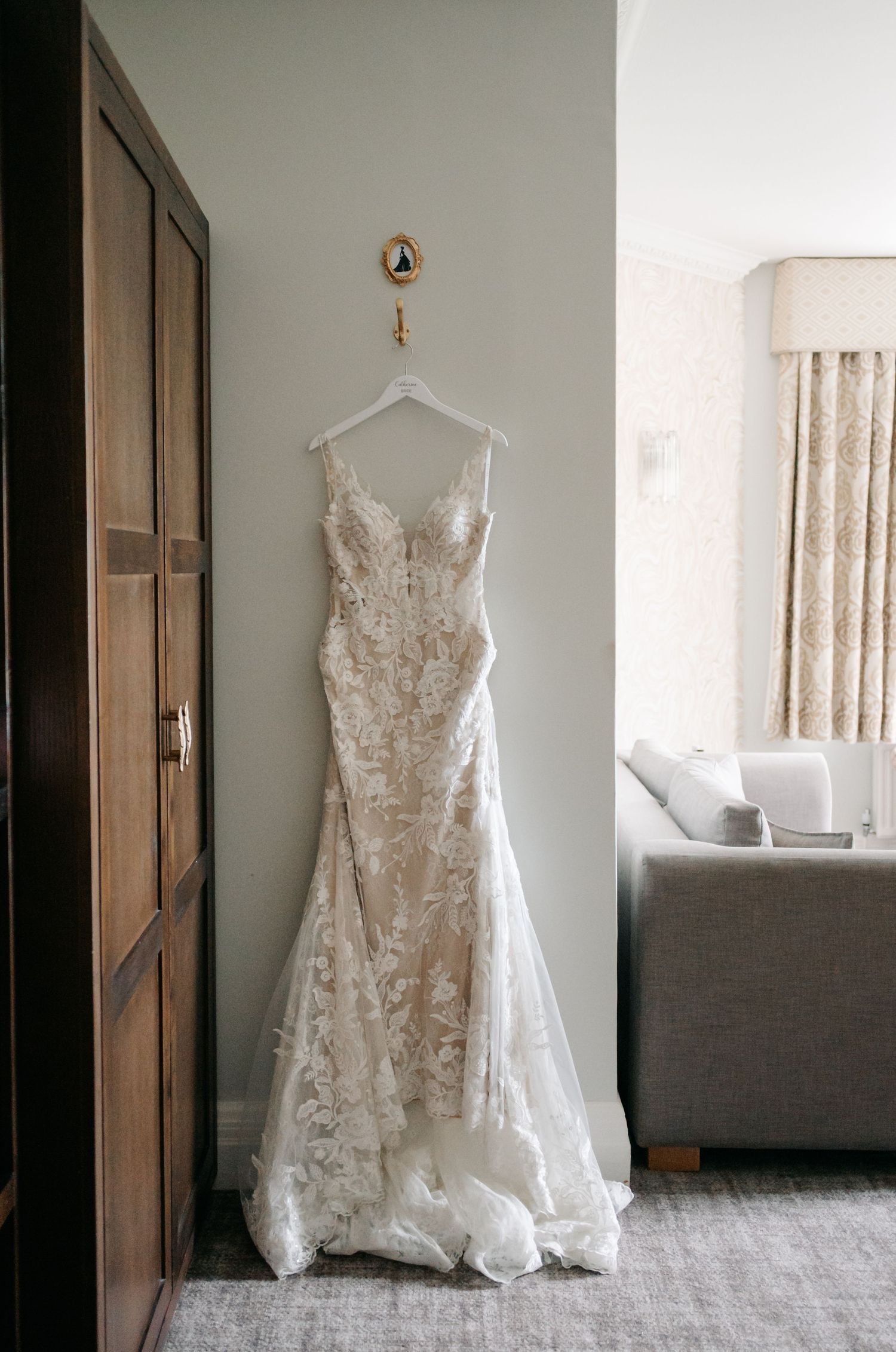 A detailed lace wedding dress hangs elegantly against a light wall, showcasing its intricate floral patterns.