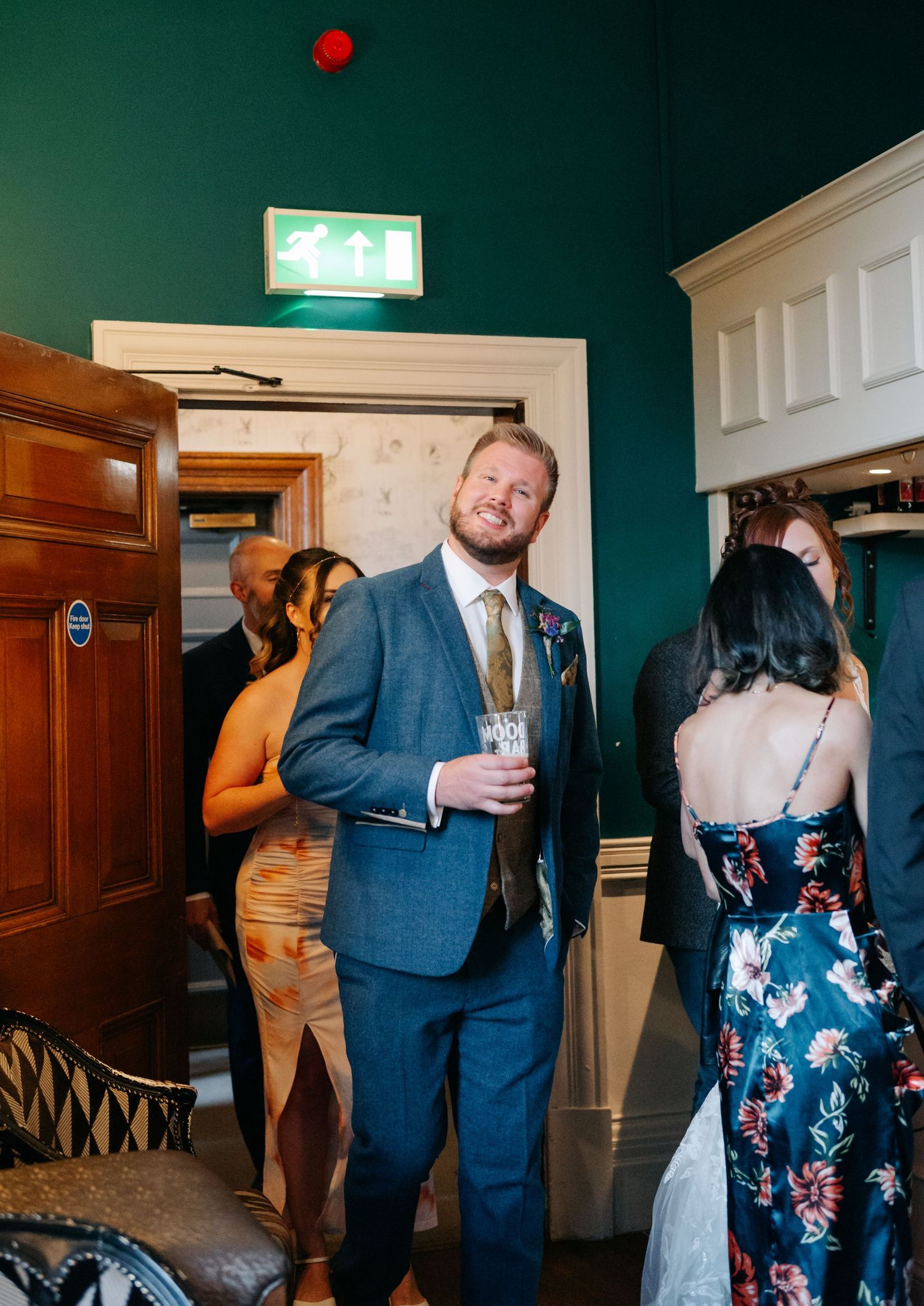 Wedding guests mingle and dance at an indoor reception with dark green walls and wooden doors.