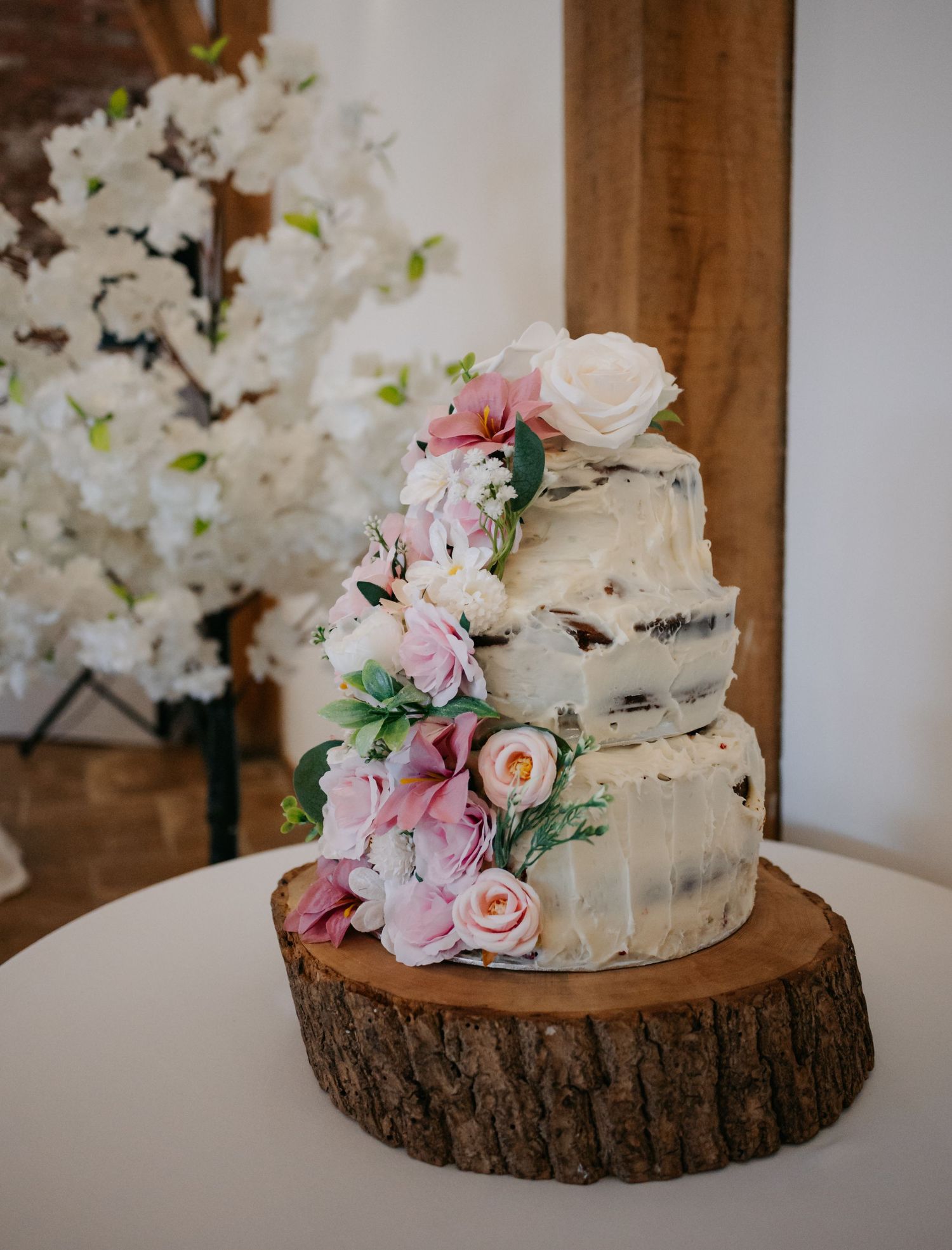 Rustic wedding cake decorated with pink and white flowers sits on wooden tree slice.