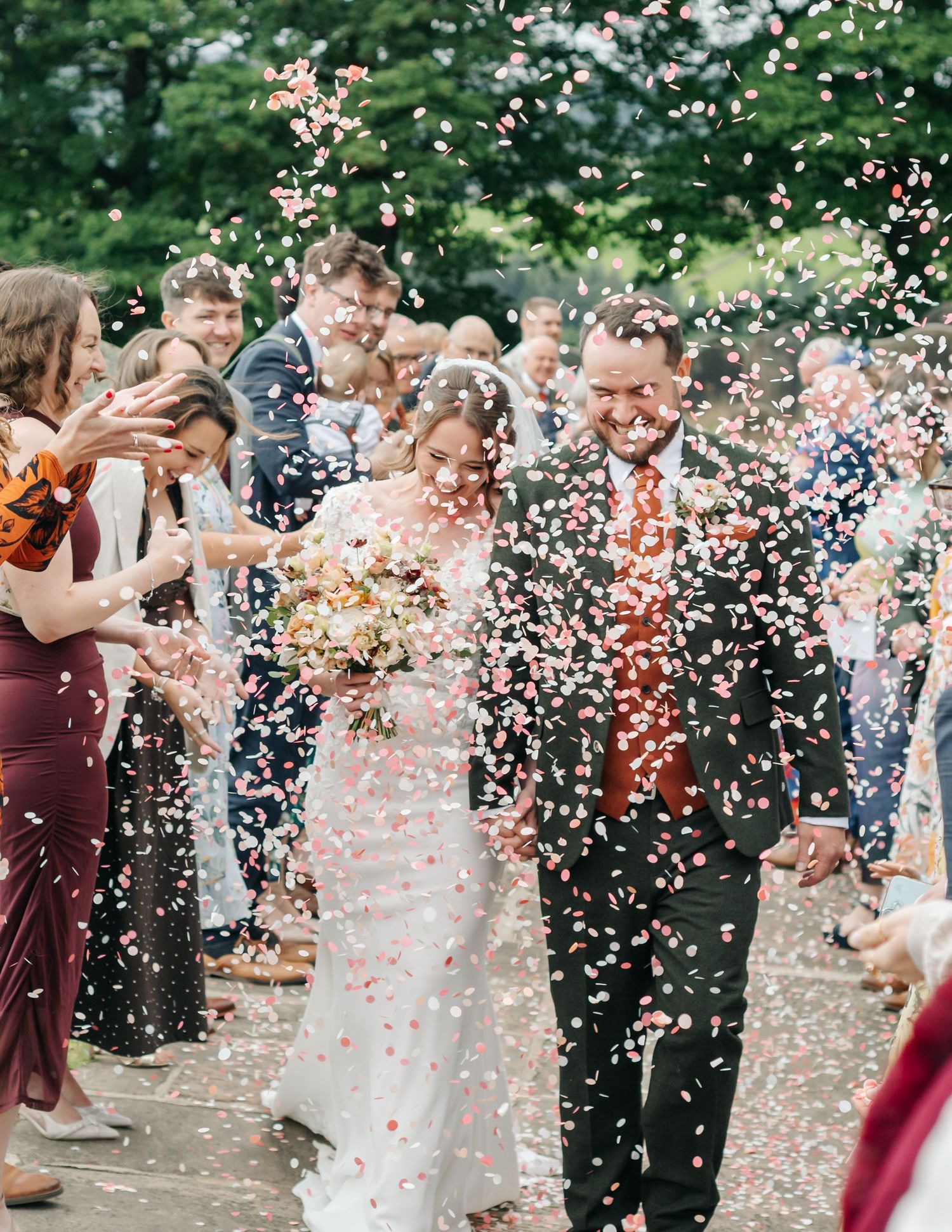 Smiling bride and groom walk hand in hand through a shower of colorful confetti as wedding guests cheer them on outdoors.