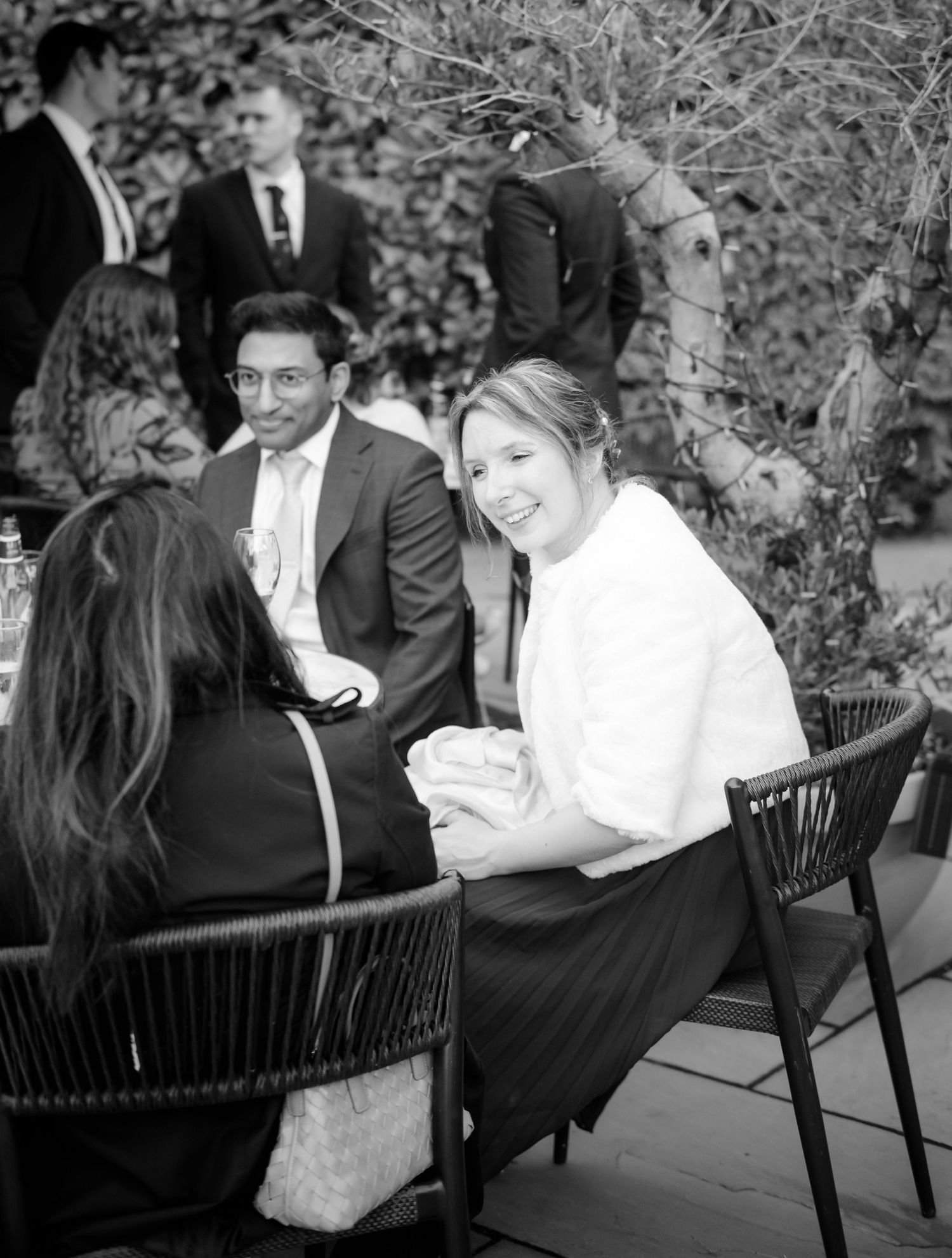 A black and white photo of smiling wedding guests chatting at an outdoor table while other guests in suits mingle in the background.