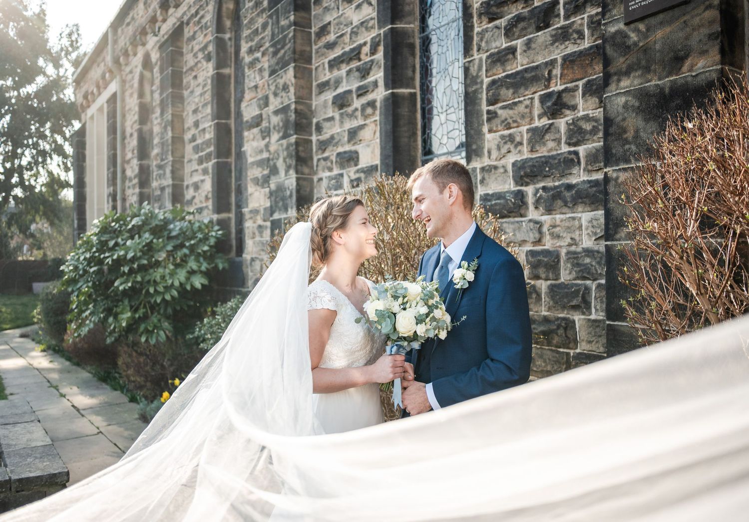 A couple shares a romantic moment outside a stone church wall while the bride's veil flows in the breeze.