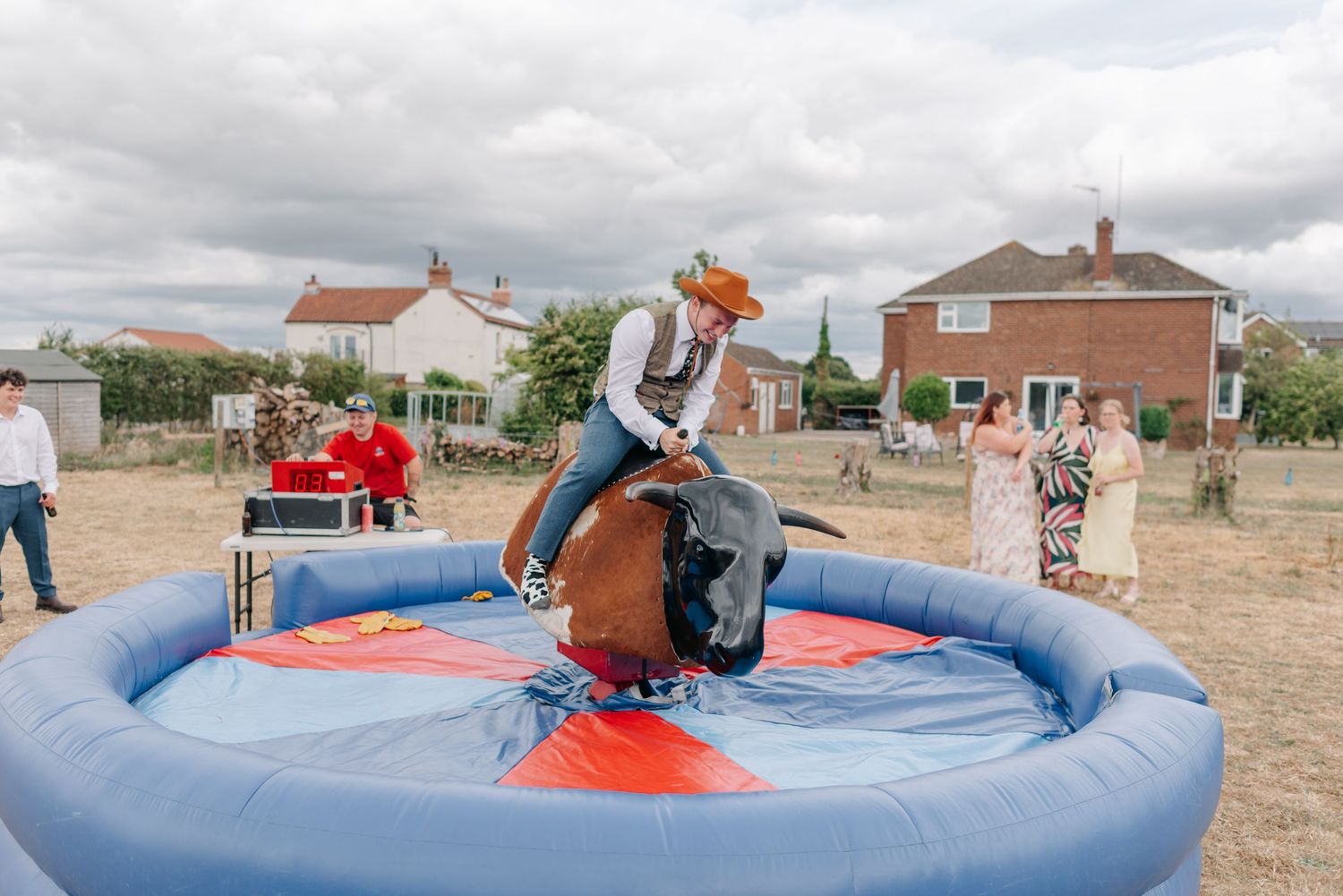 People enjoying a mechanical bull ride at an outdoor wedding reception with red and blue inflatable mat.
