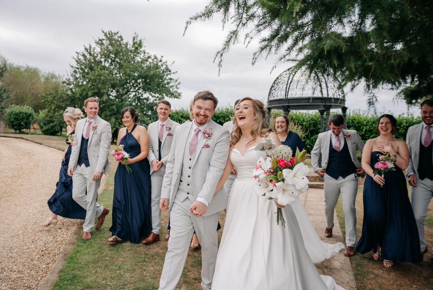 Wedding party in navy and grey attire walking together on a gravel path outdoors.