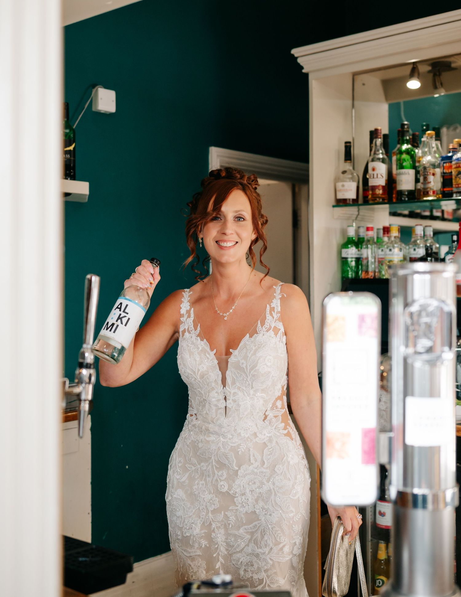 A bride in white lace dress poses at a bar with bottles and glassware.