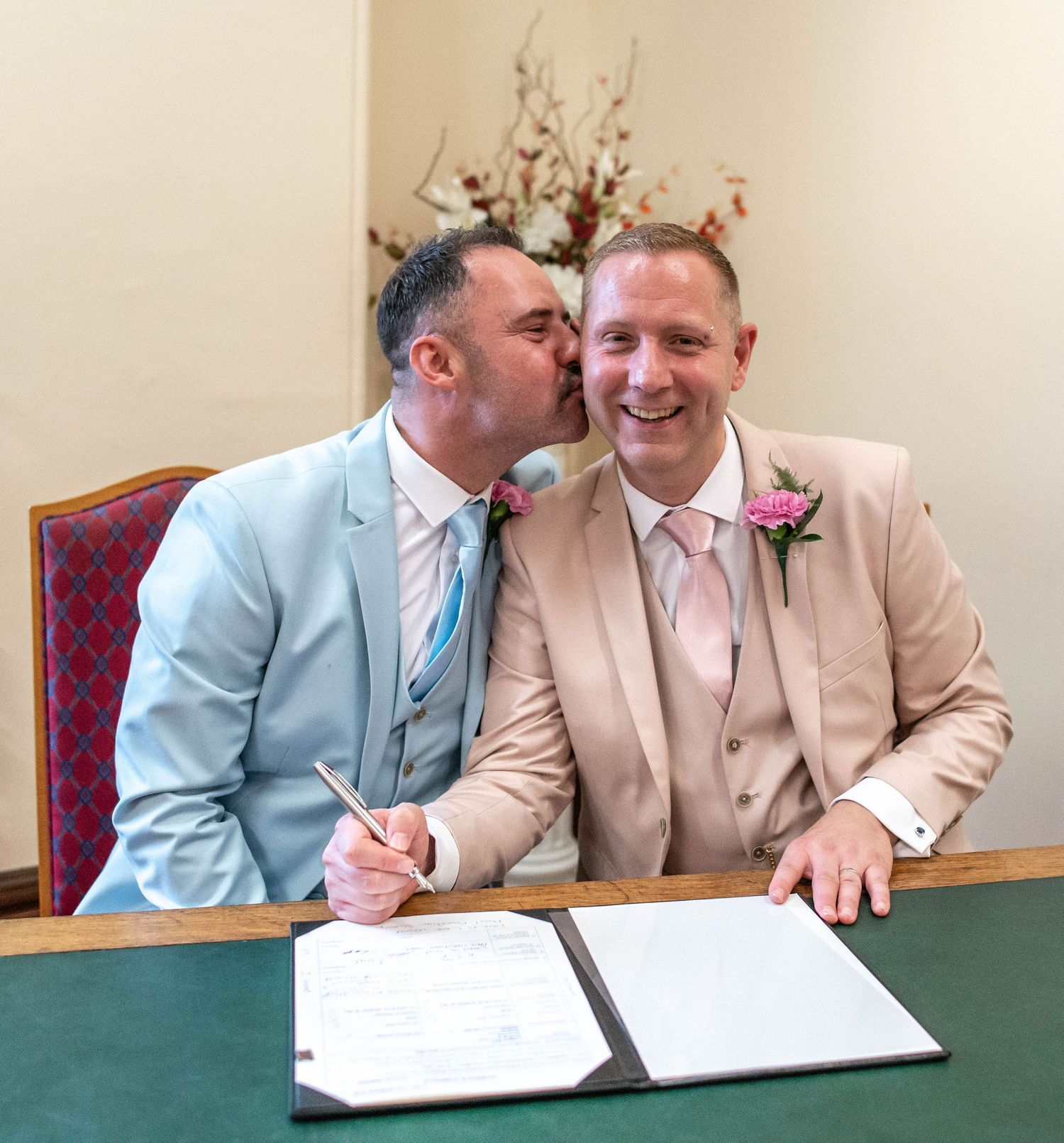 Happy couple in pastel suits share tender moment while signing marriage documents.