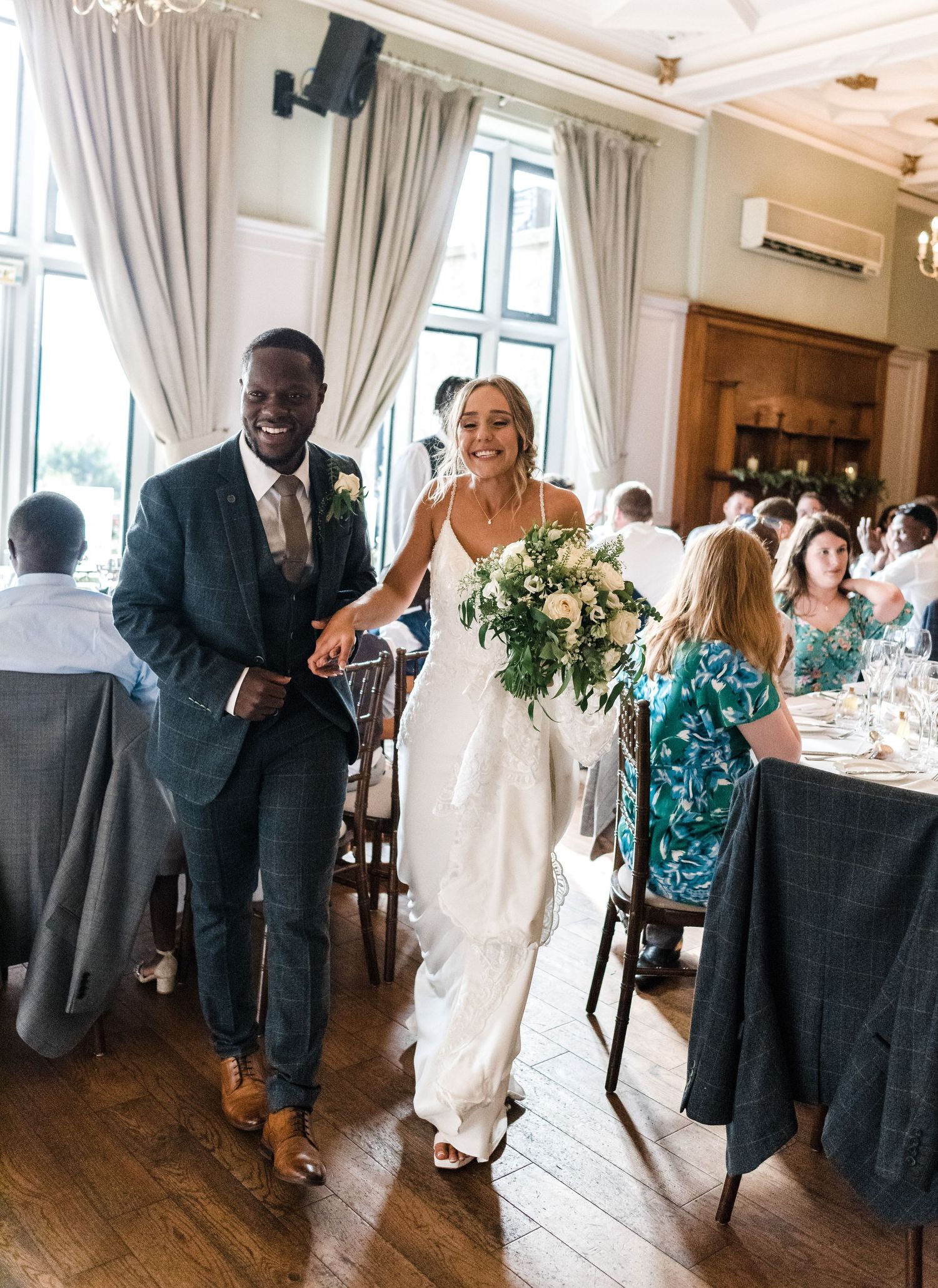 Wedding celebration in an elegant reception hall with guests seated at tables with grey linens and floral centerpieces.