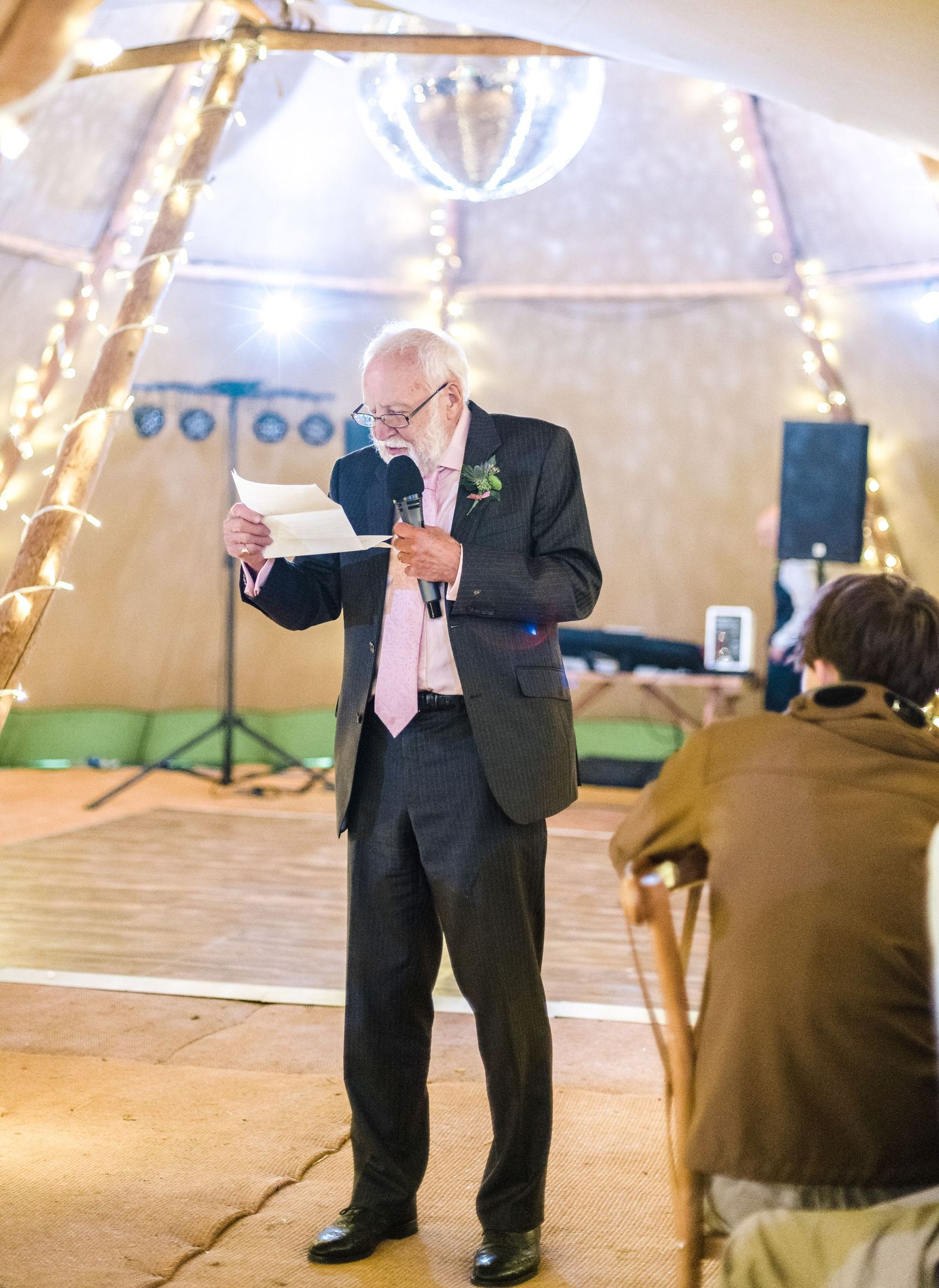 A speaker giving a speech at a wedding reception in a tent venue with string lights.