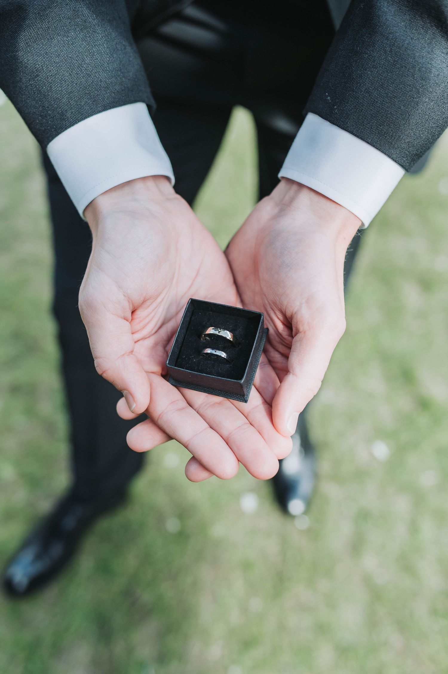 Close-up of hands holding a black ring box with two wedding bands against formal attire.