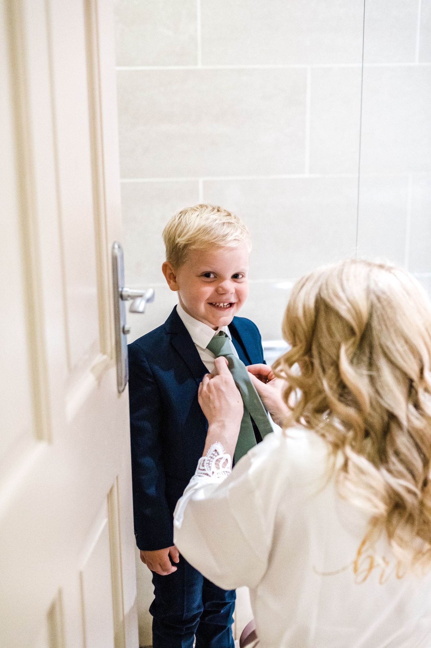 A young ring bearer in a navy suit and tie gets help adjusting his outfit before a wedding ceremony.