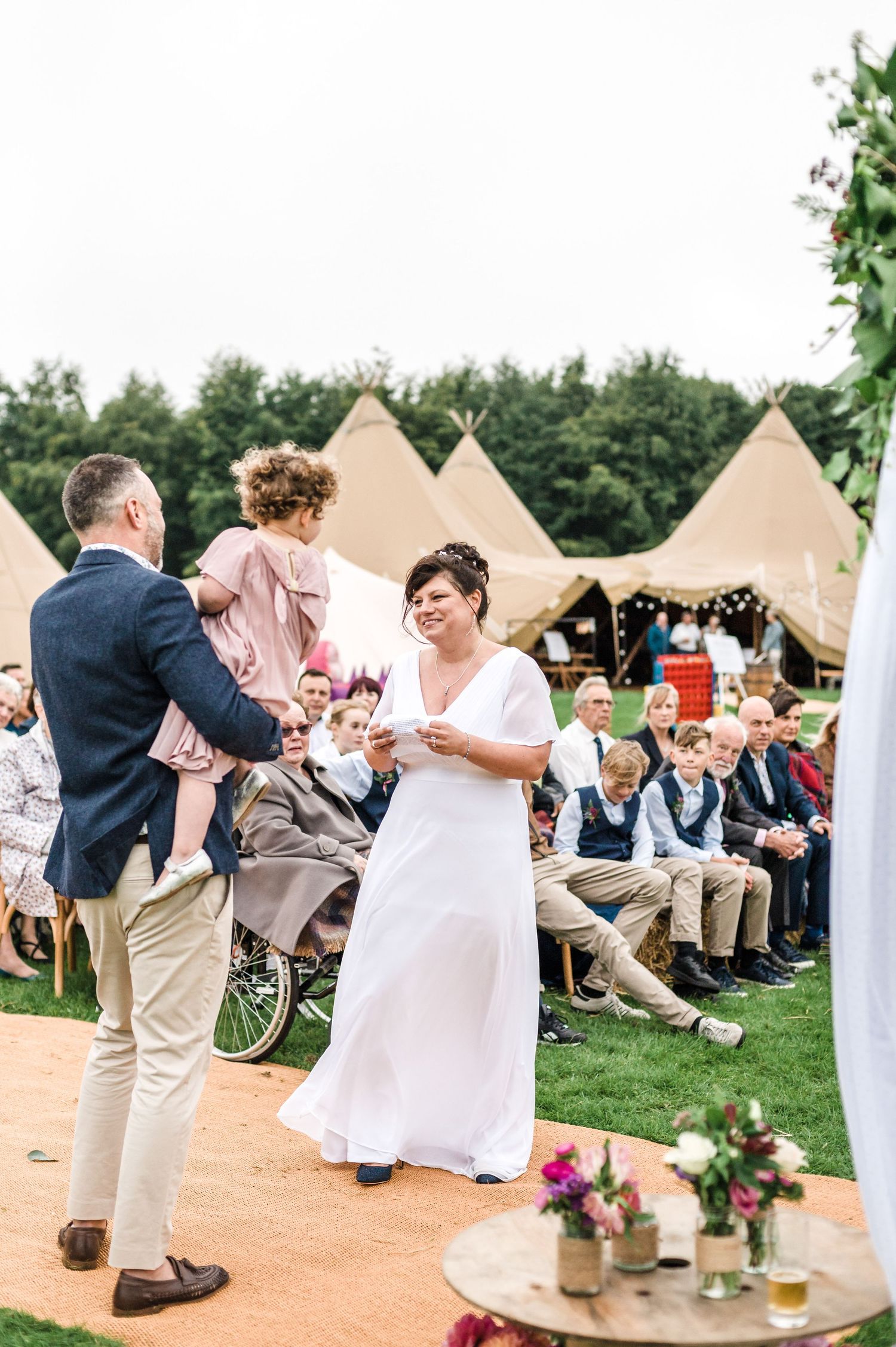 Outdoor wedding ceremony with guests seated on lawn chairs and teepee tents in the background.