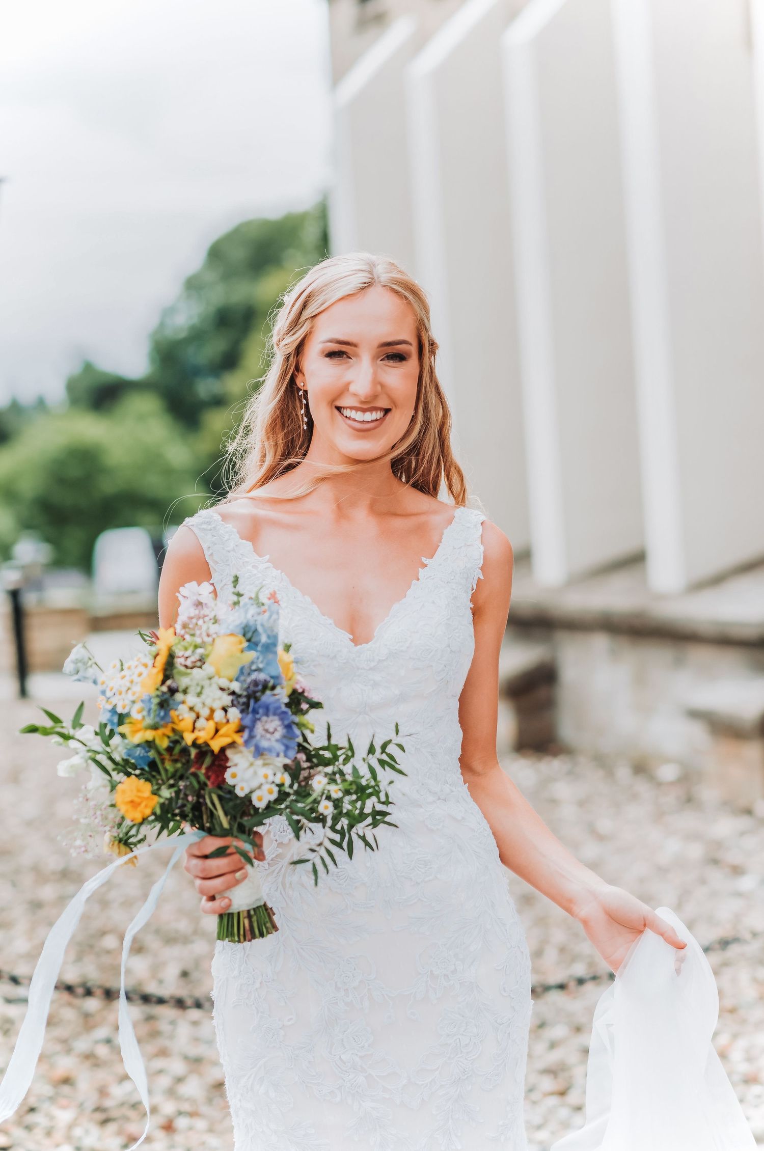 Summer bride in lace V-neck dress holding colorful wildflower bouquet outside white pillared building.