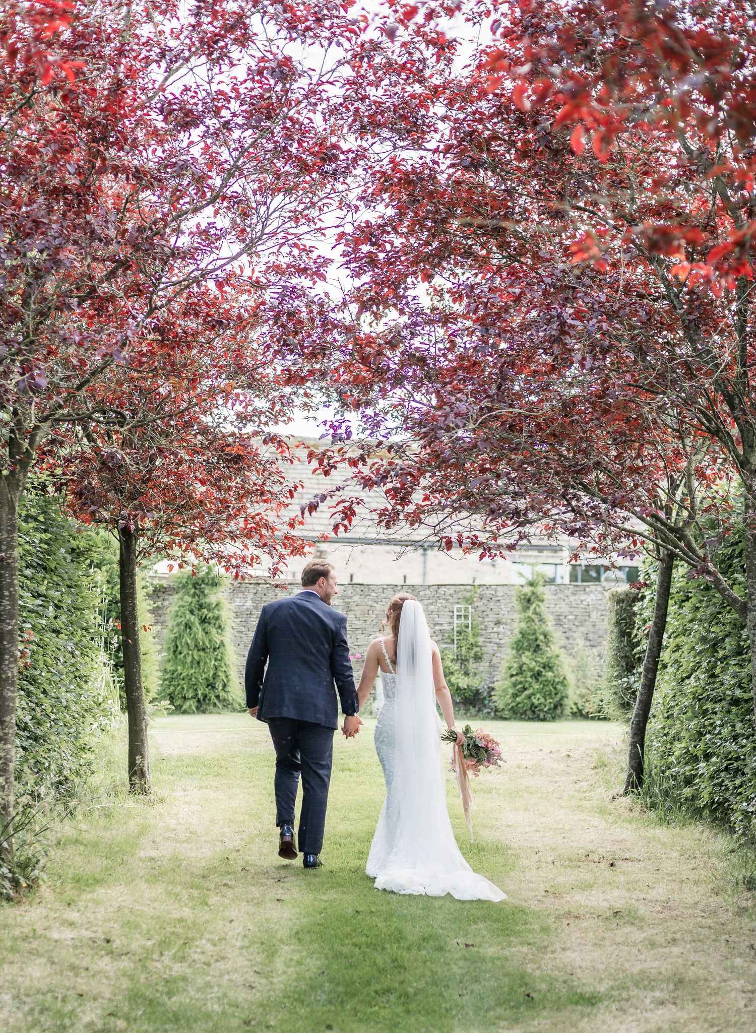 Couple walking down garden path under canopy of red-leafed trees.