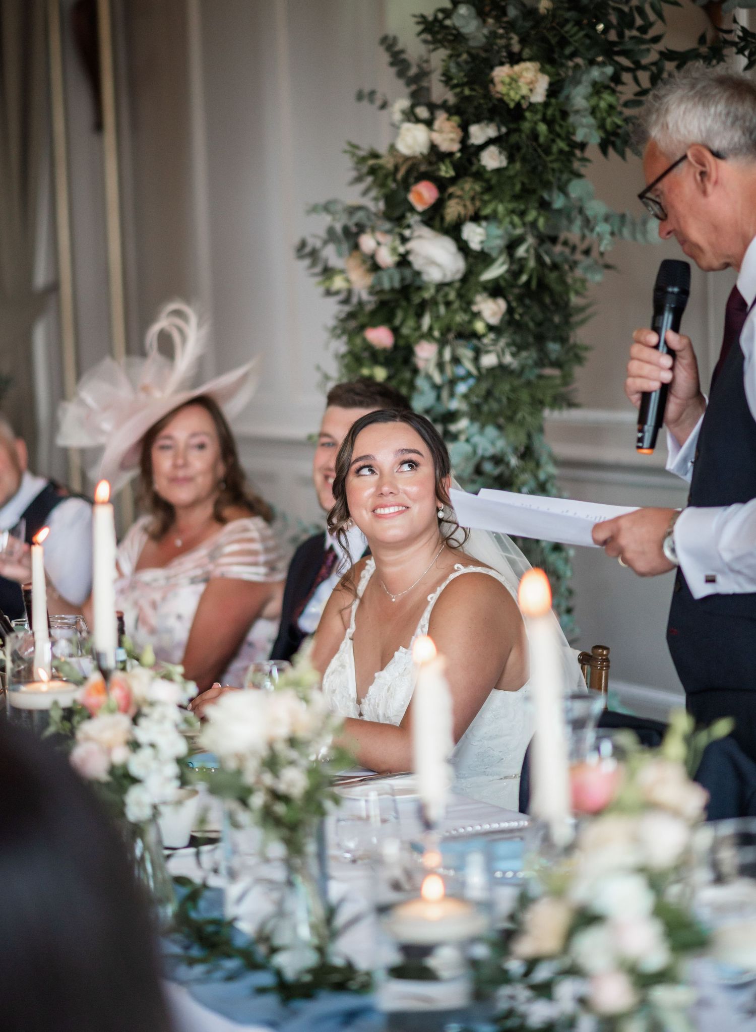 An elegant wedding reception with floral centerpieces and a speaker addressing guests.