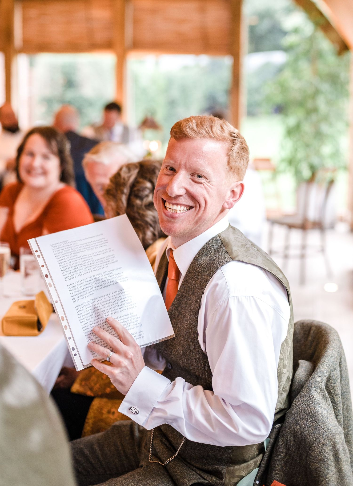 Guest in tweed vest smiles while holding papers during wedding reception.