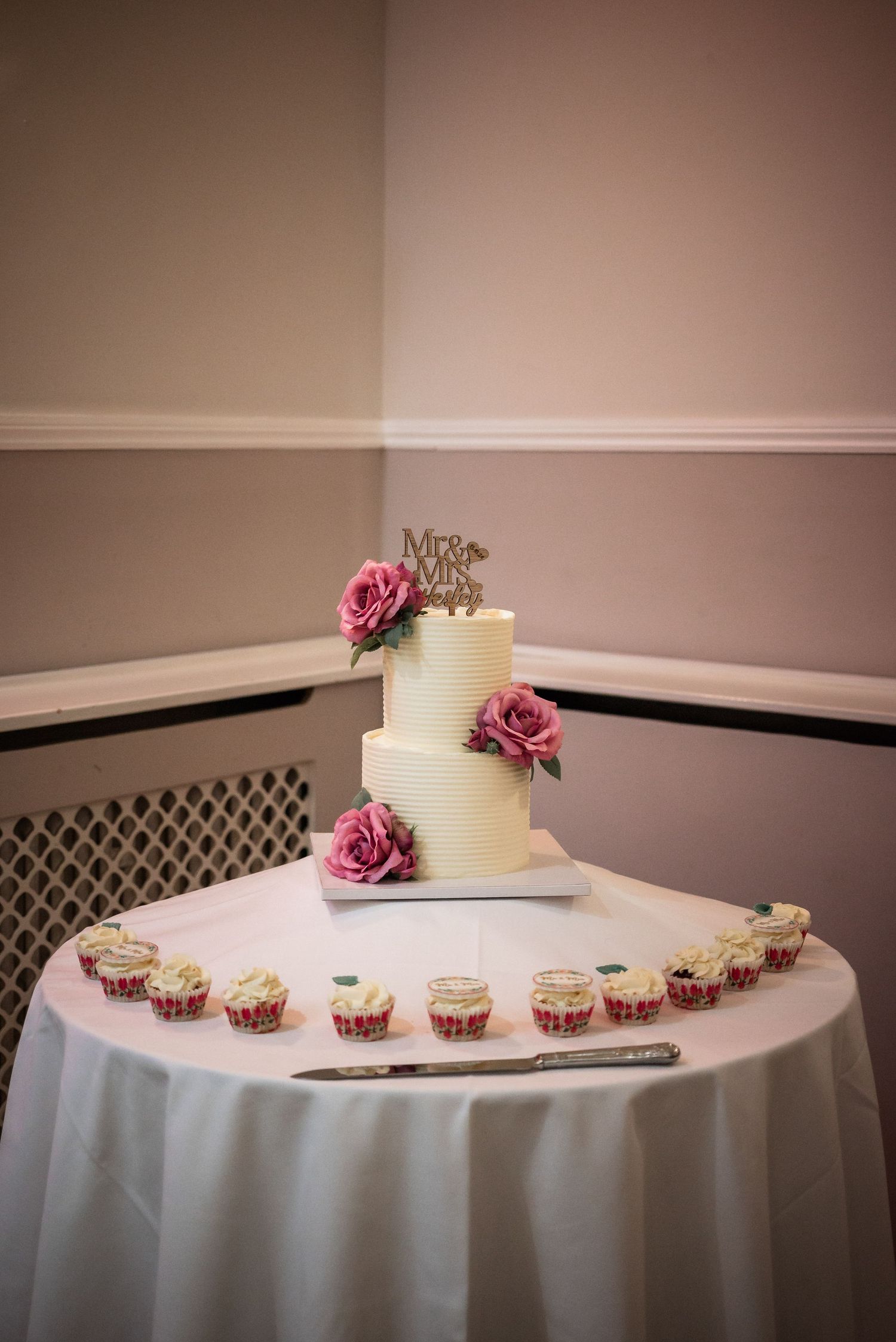 White two-tier wedding cake with pink roses and cupcakes displayed on round table with white tablecloth.