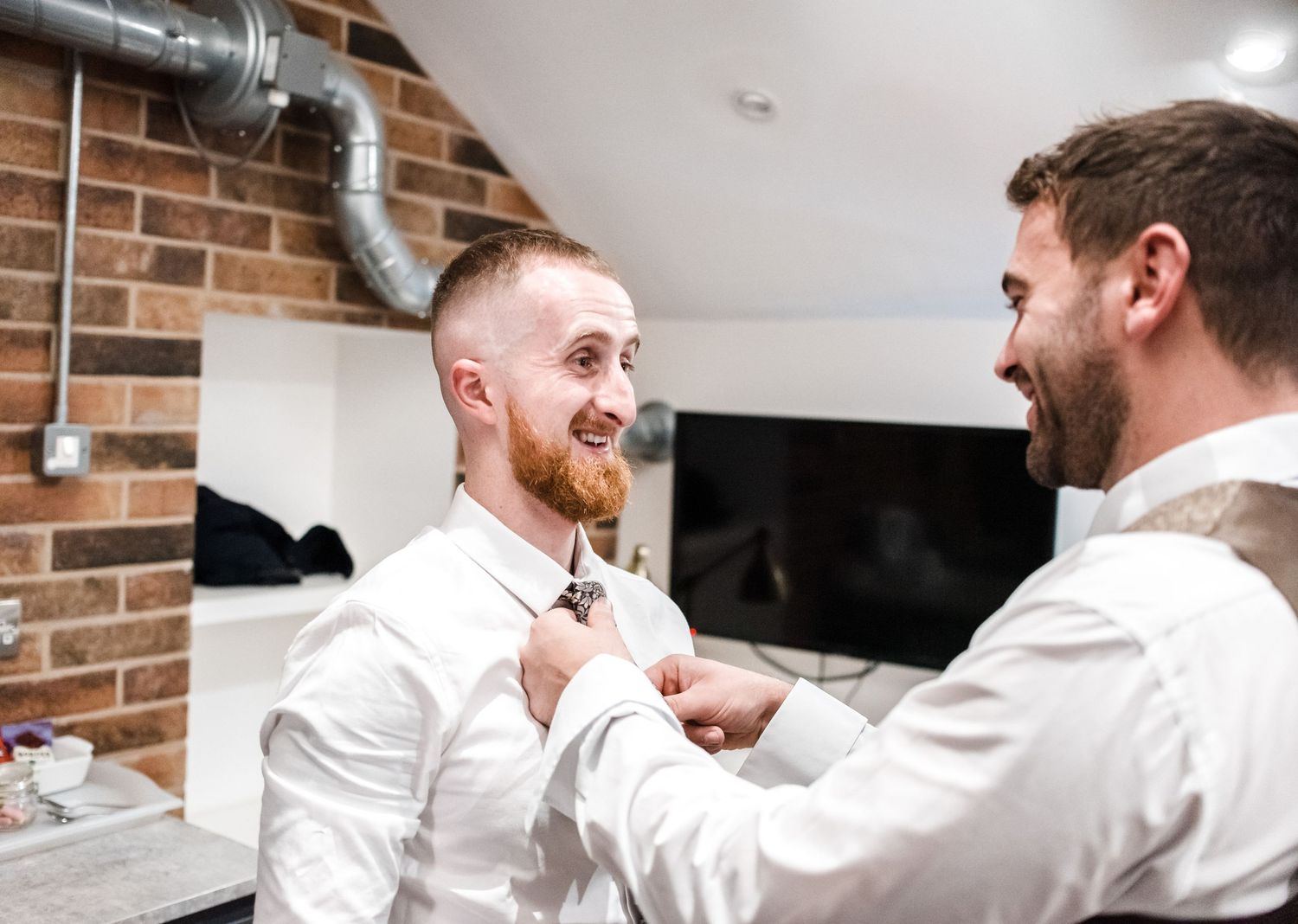 Groomsman helping adjust another groomsman's tie in brick-walled room.