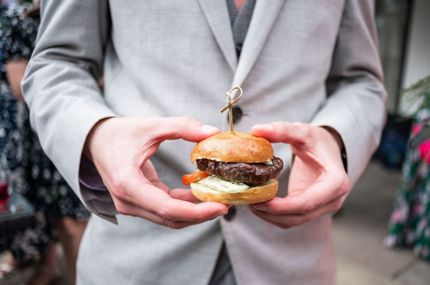 Person in grey suit holding a gourmet burger at wedding reception.