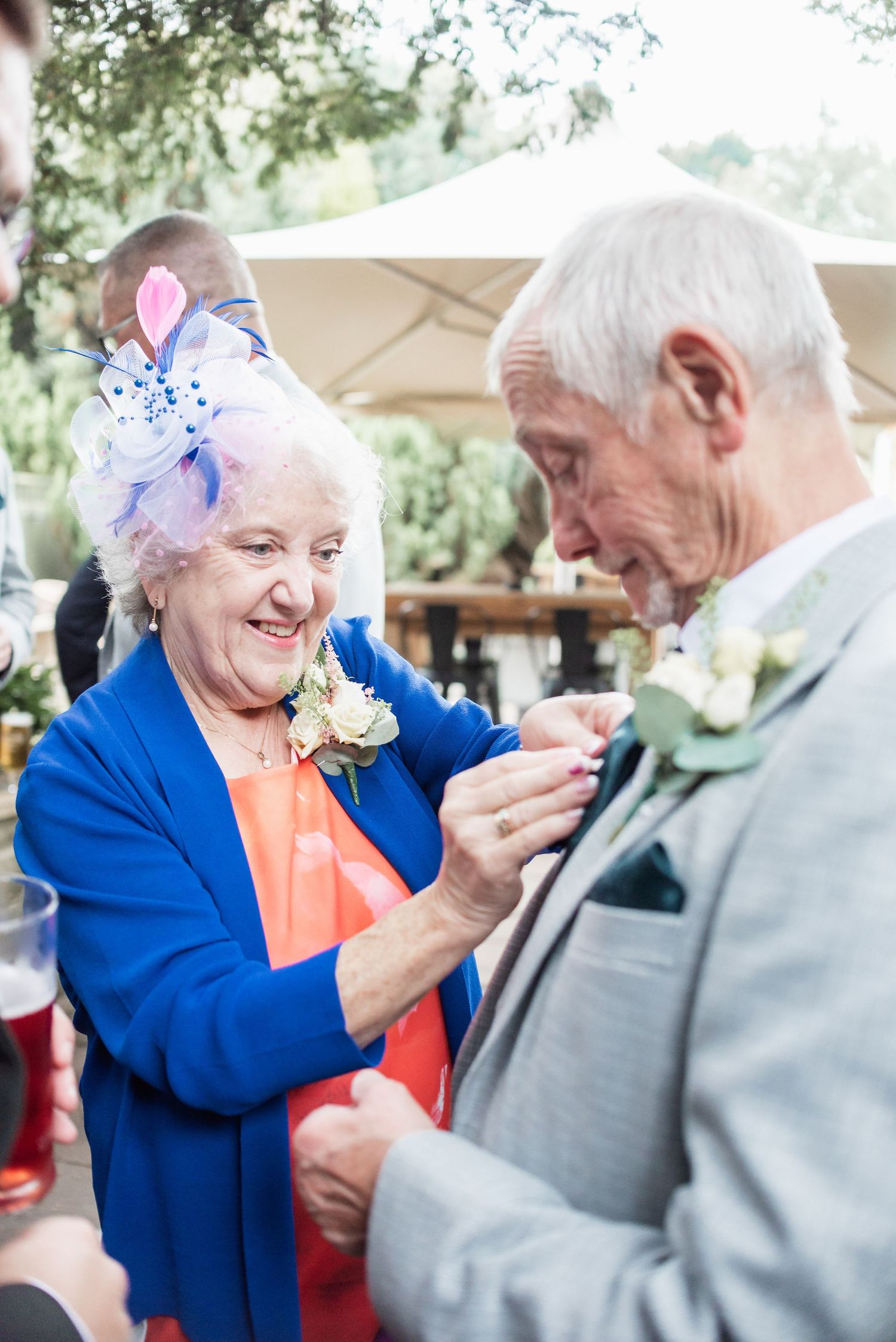 Elderly couple in formal attire share sweet moment pinning boutonniere at outdoor event.