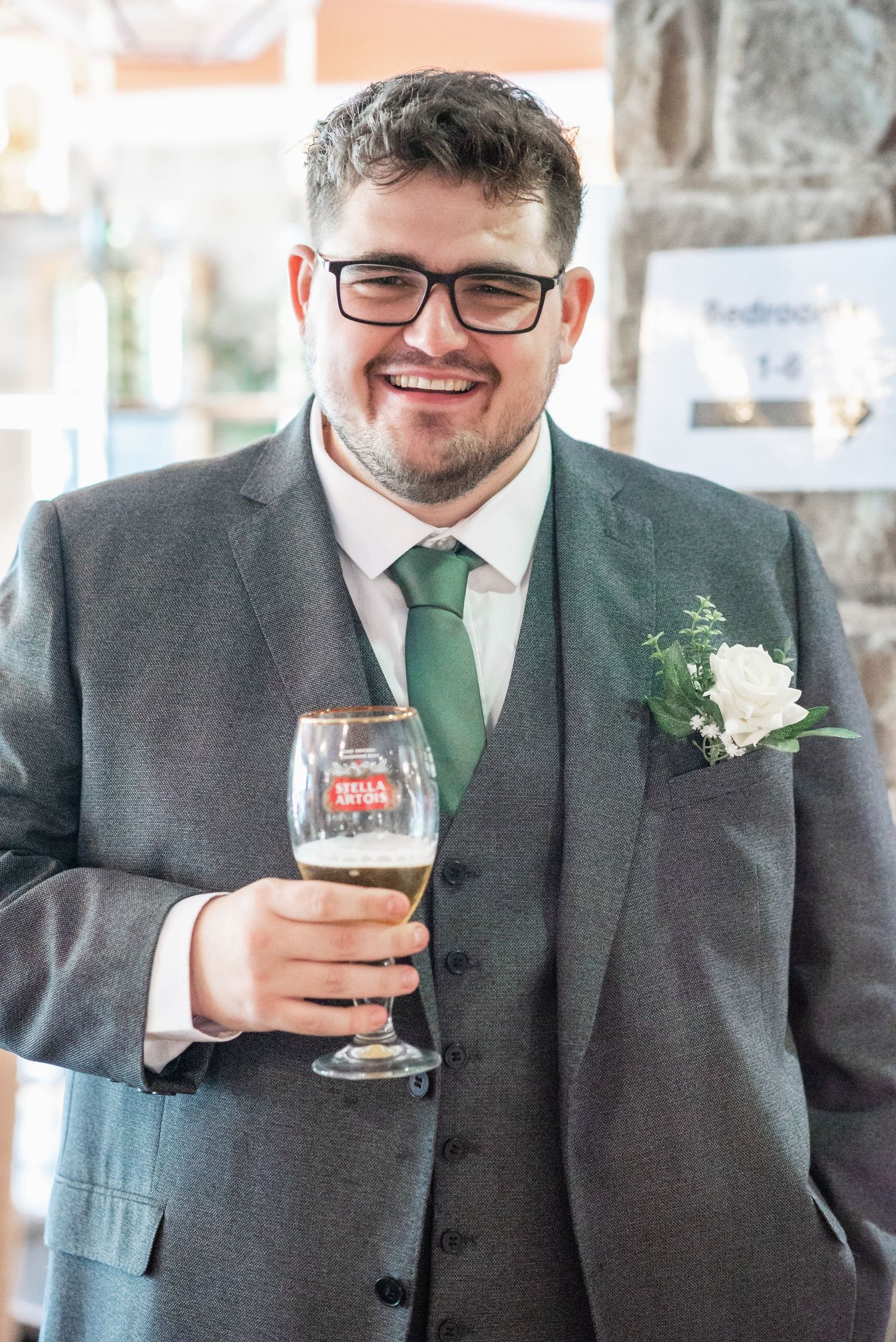 Wedding guest in grey suit and green tie holding beer glass at reception.