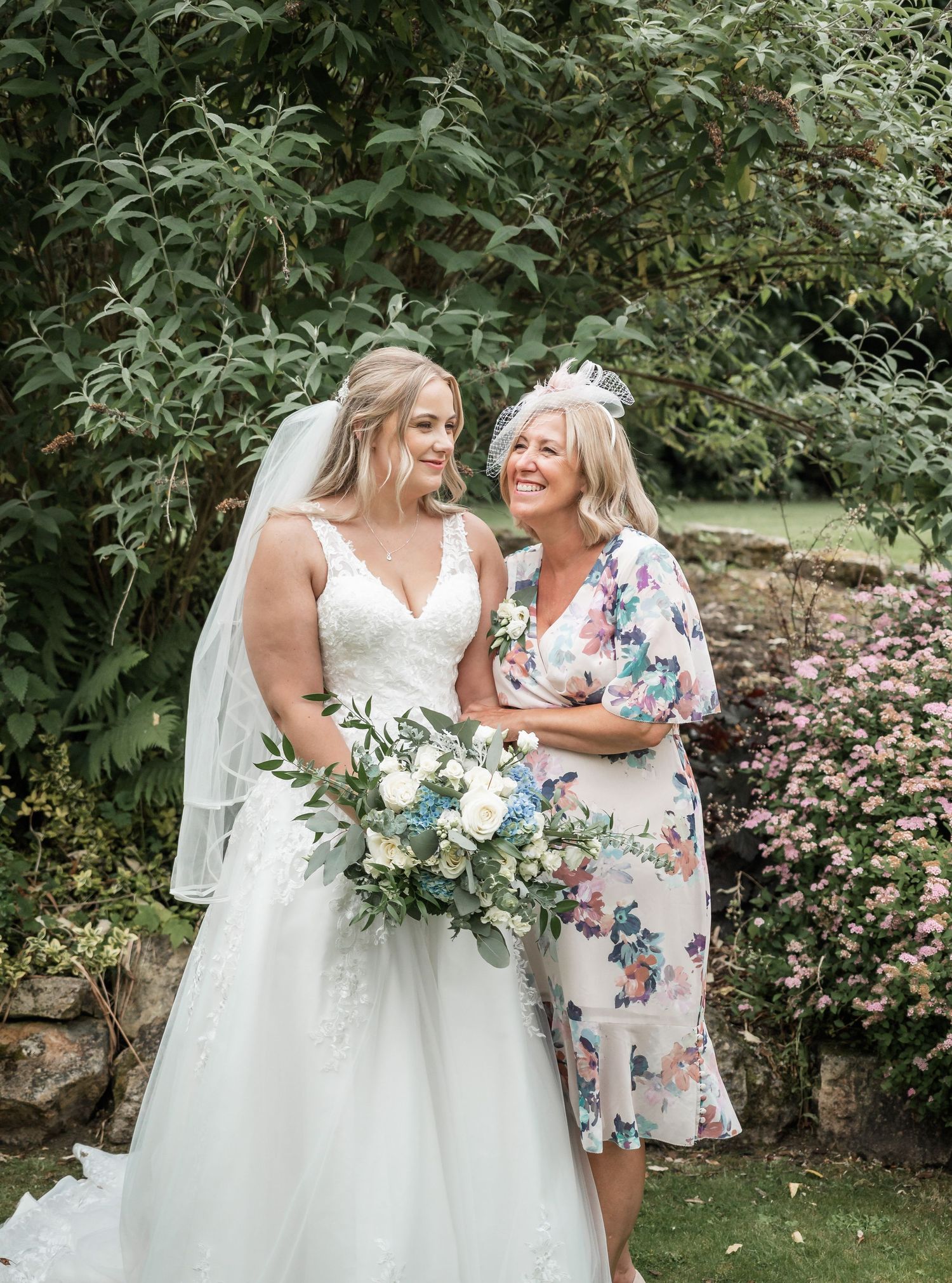 A bride in white holds a bouquet while sharing a joyful moment in a garden setting.