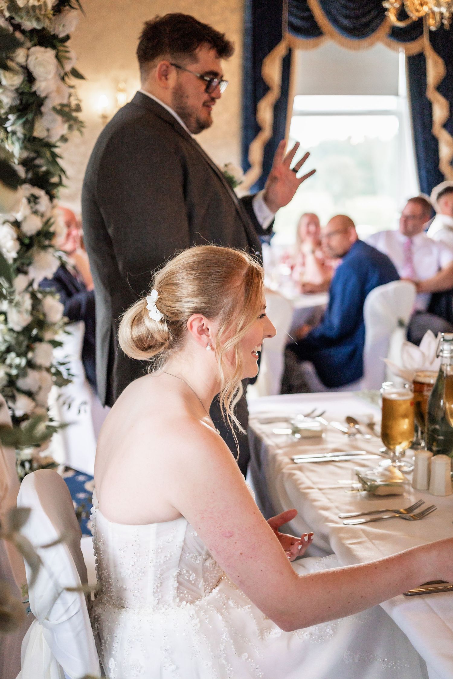 Candid moment during wedding speeches with guests seated in elegant reception room with floral decor.