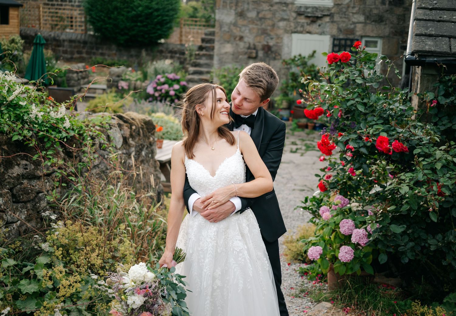 A bride and groom embrace in a romantic garden setting with red roses, the groom in a black tuxedo kissing her cheek.