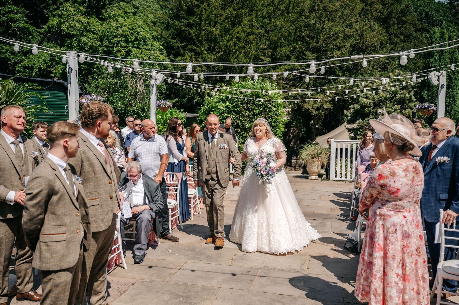 Bride walks down outdoor aisle with escort under fairy lights, guests watching at sunny garden wedding.