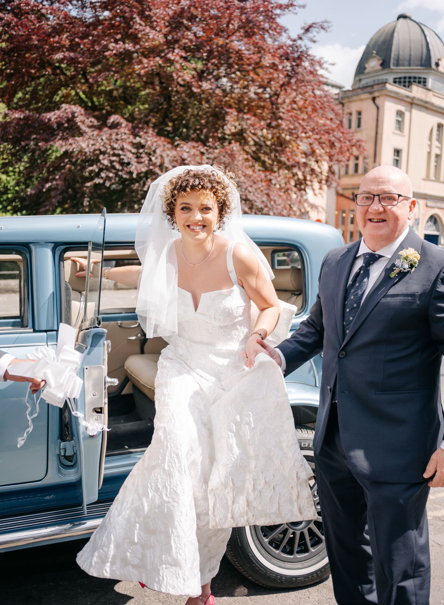 Wedding party posing with vintage blue car under pink blossoming trees.
