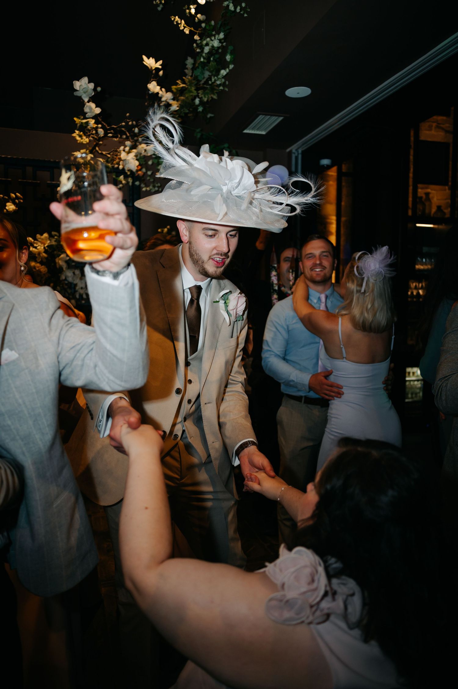 A guest wearing a large decorative white hat dances enthusiastically at a wedding reception.