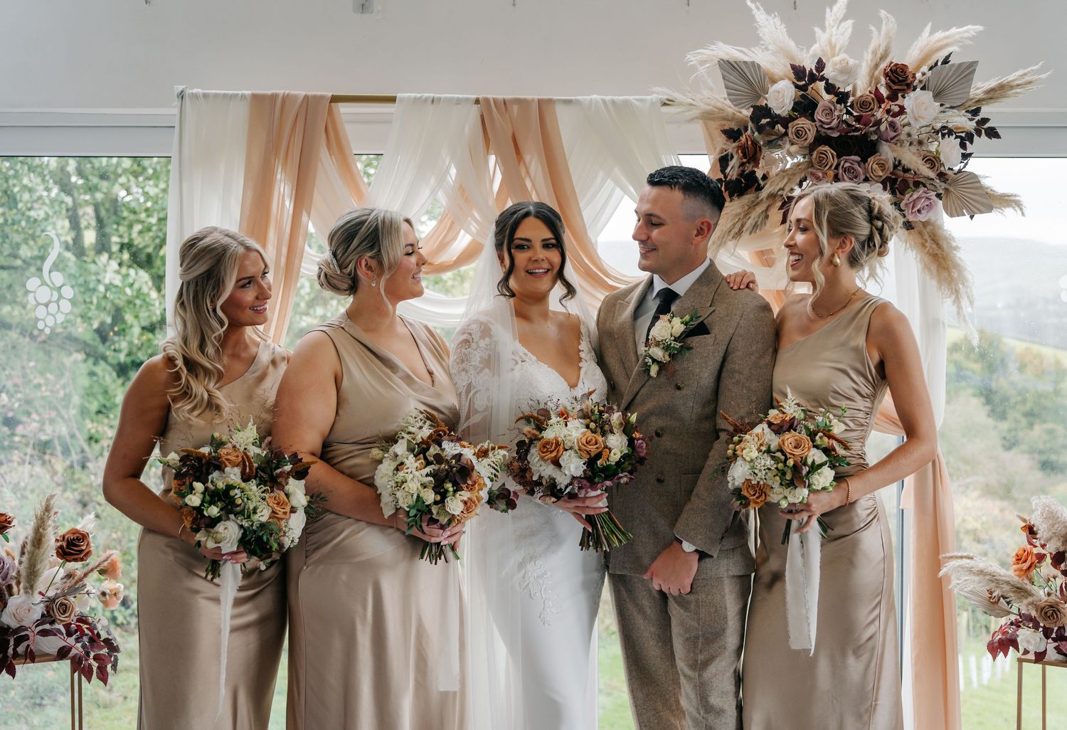Bridal party in champagne dresses holding floral bouquets against bright windows.