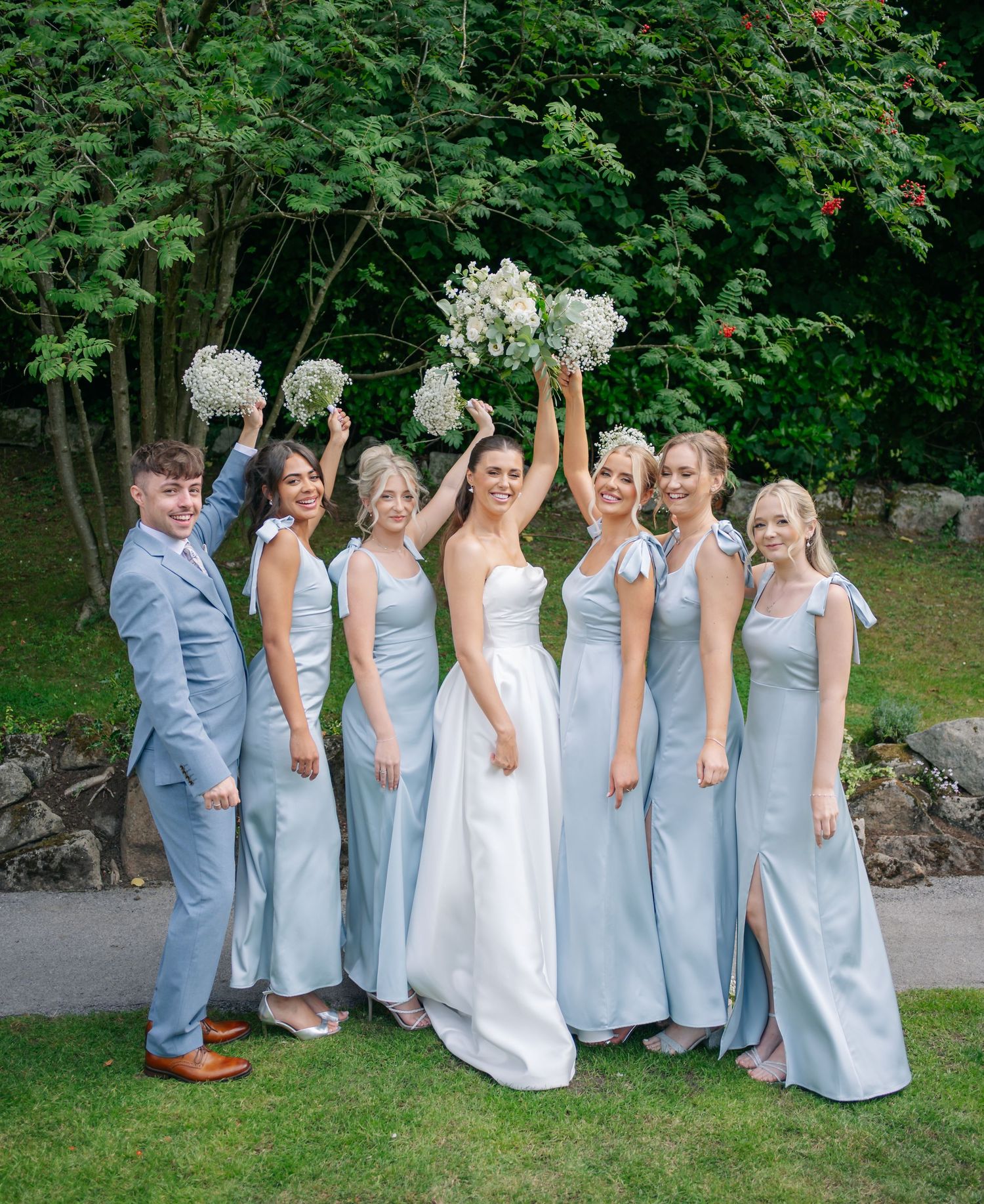 A bridal party in light blue dresses poses with bouquets on a garden path.