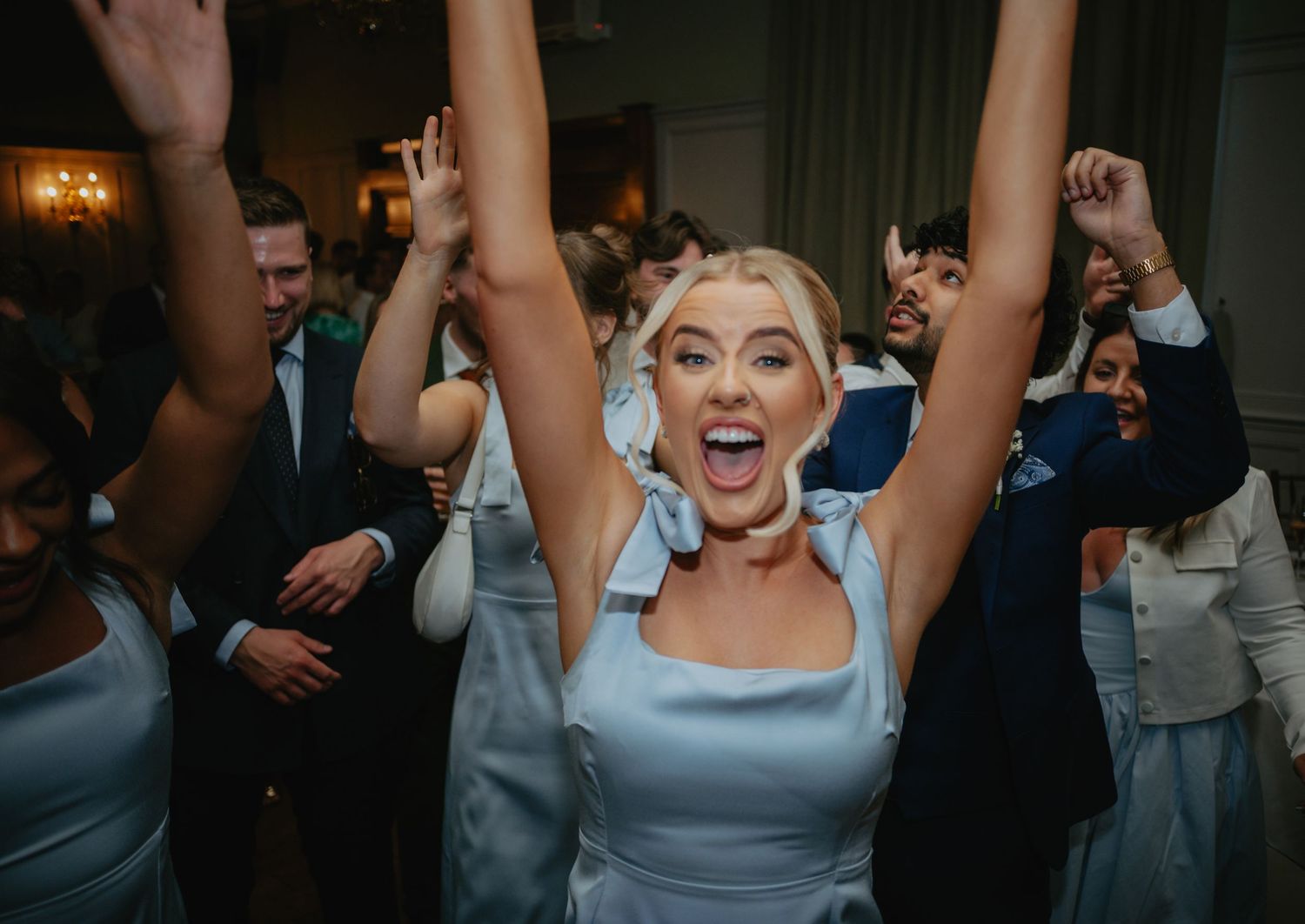 Wedding party members dancing enthusiastically with raised arms in a dimly lit reception venue.