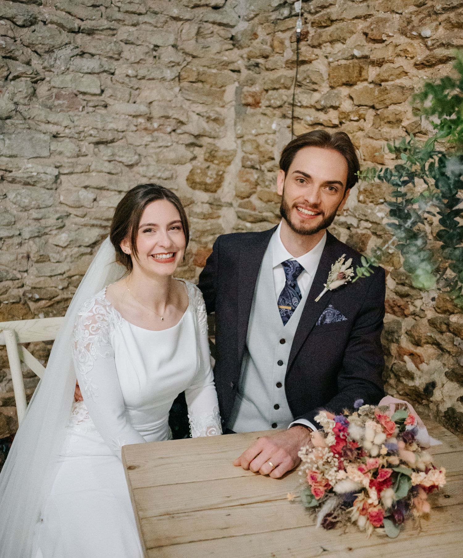 A wedding couple sits together at a rustic stone wall venue with a colorful floral bouquet on the wooden table.