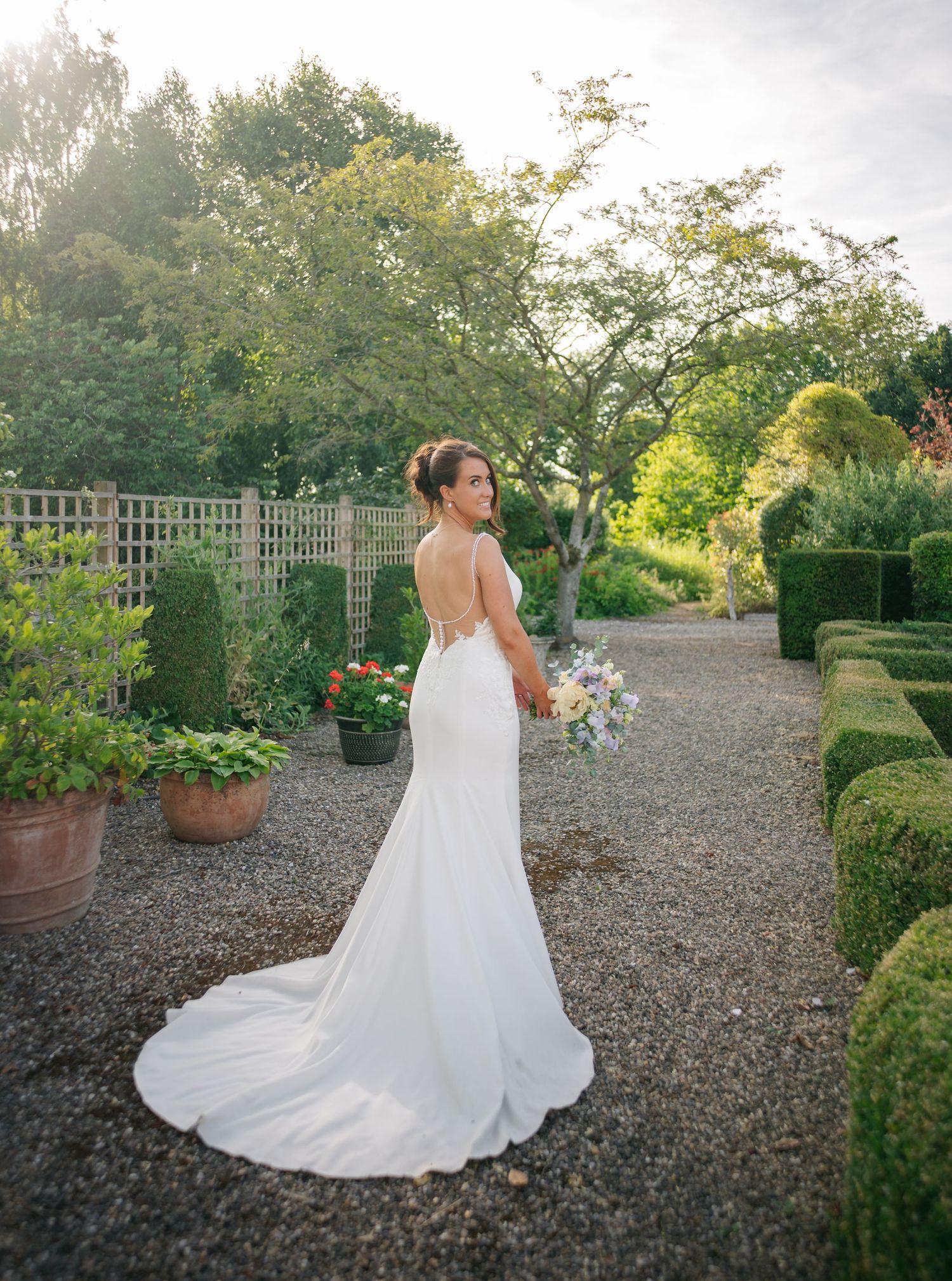 Elegant bride in backless white gown holds bouquet while standing in formal garden with gravel path.