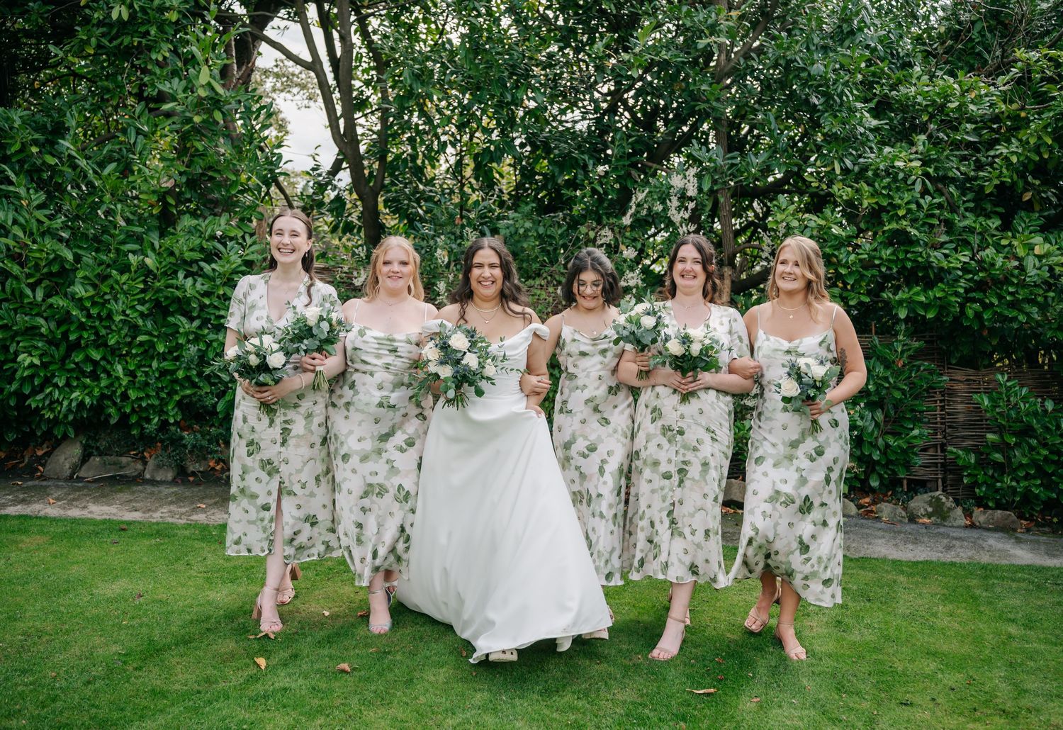 Wedding party in floral dresses poses together on a green lawn during an outdoor celebration.