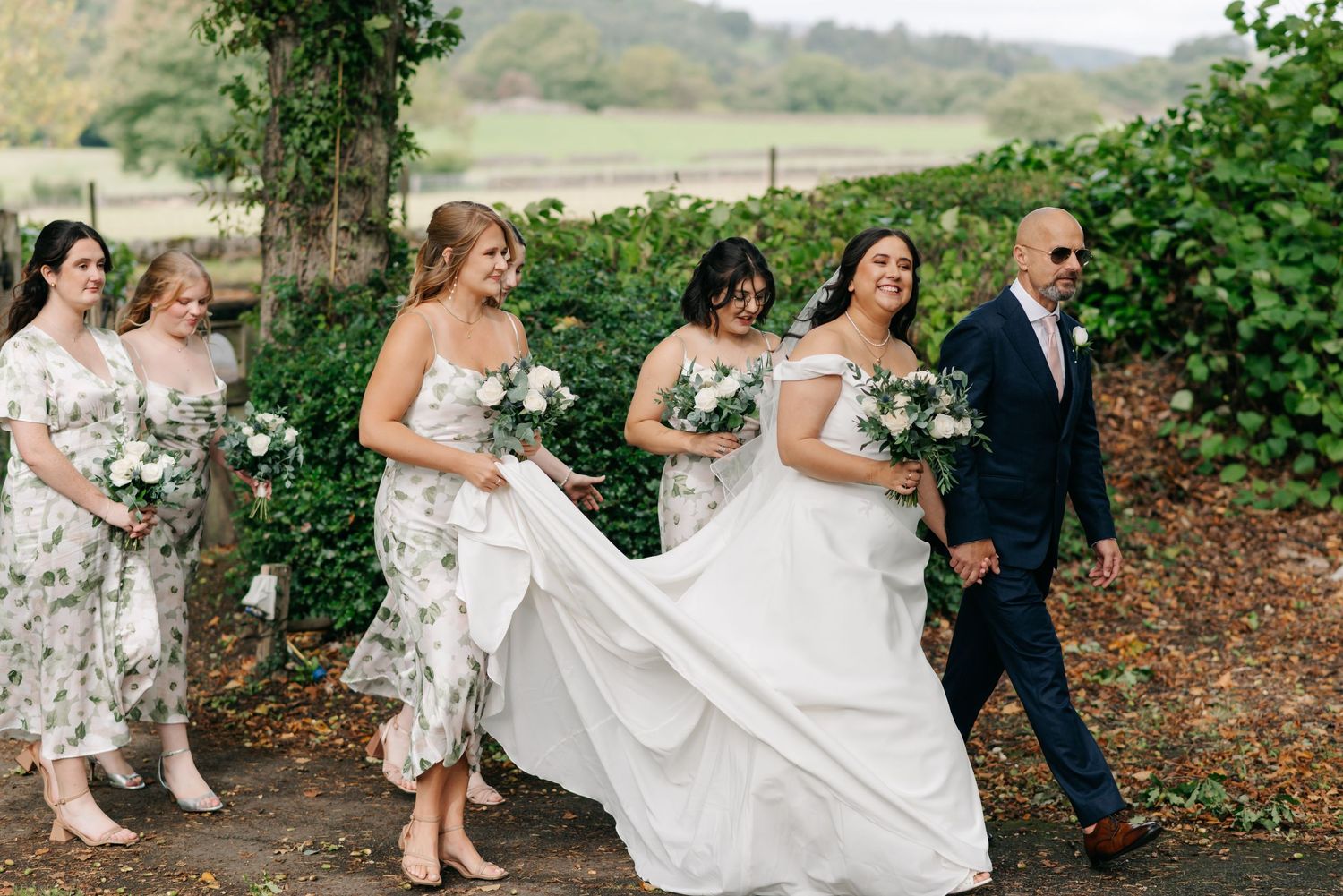 Wedding party walking through a lush green garden path with flowing dresses and veils.