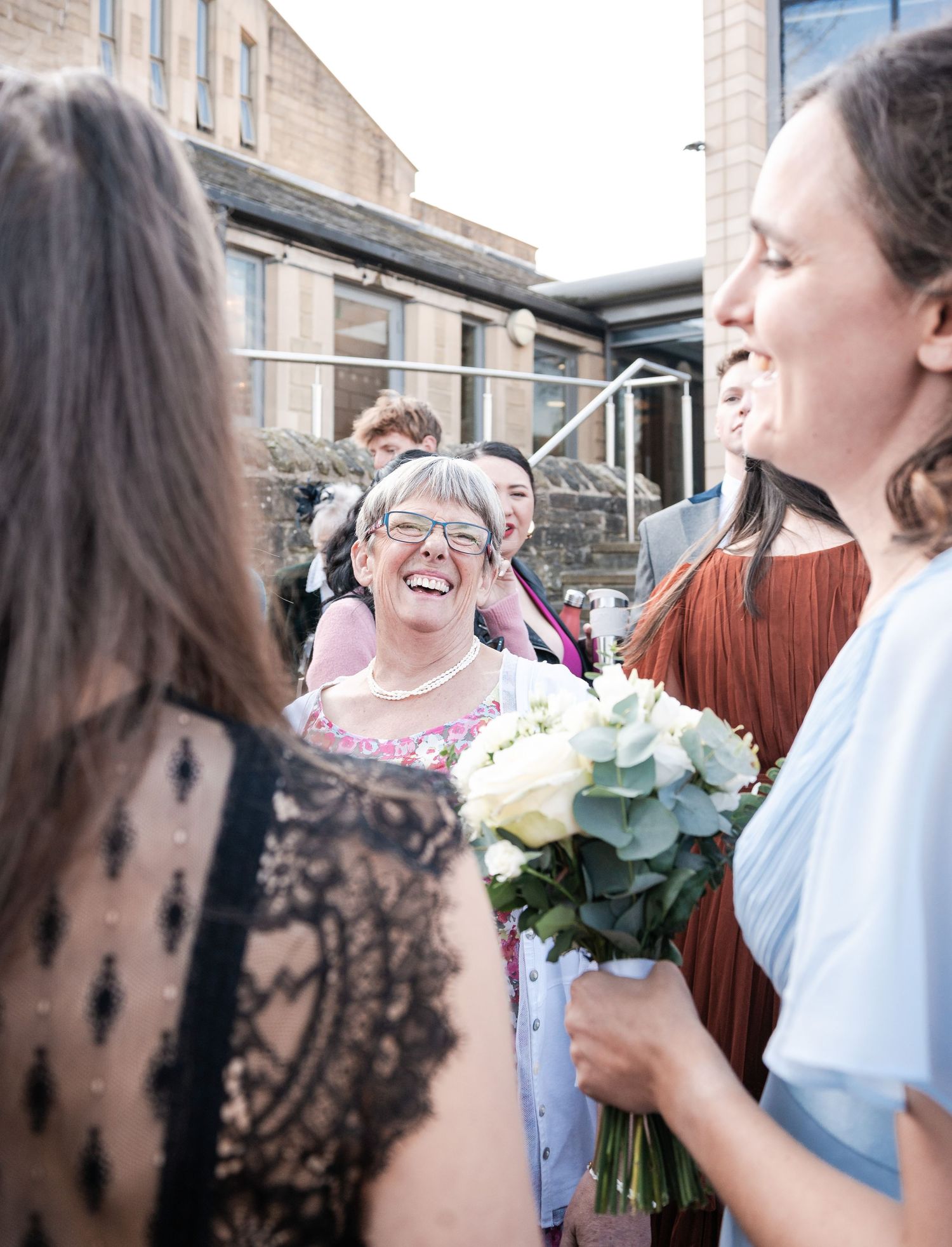 Wedding party members share joyful laughter while holding bouquets during an indoor celebration.