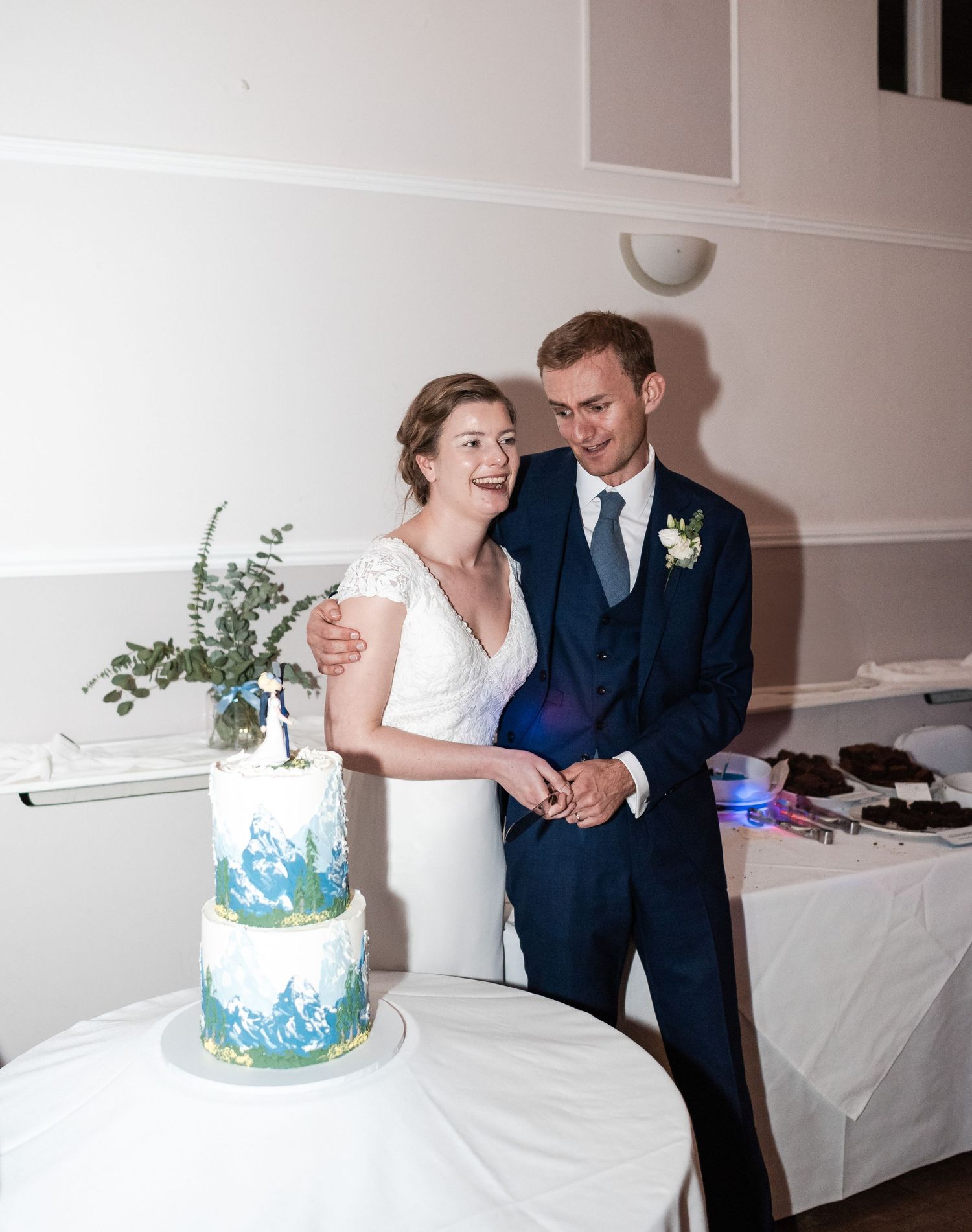 A couple stands together next to their blue and white watercolor wedding cake at their reception.