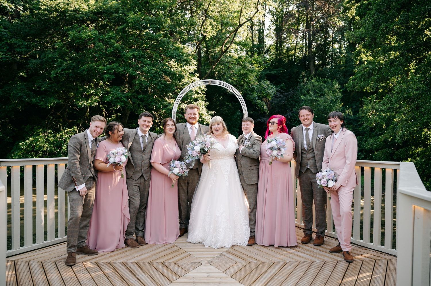 Wedding party in dusty rose and grey attire posing together on a wooden deck with a circular arch backdrop.