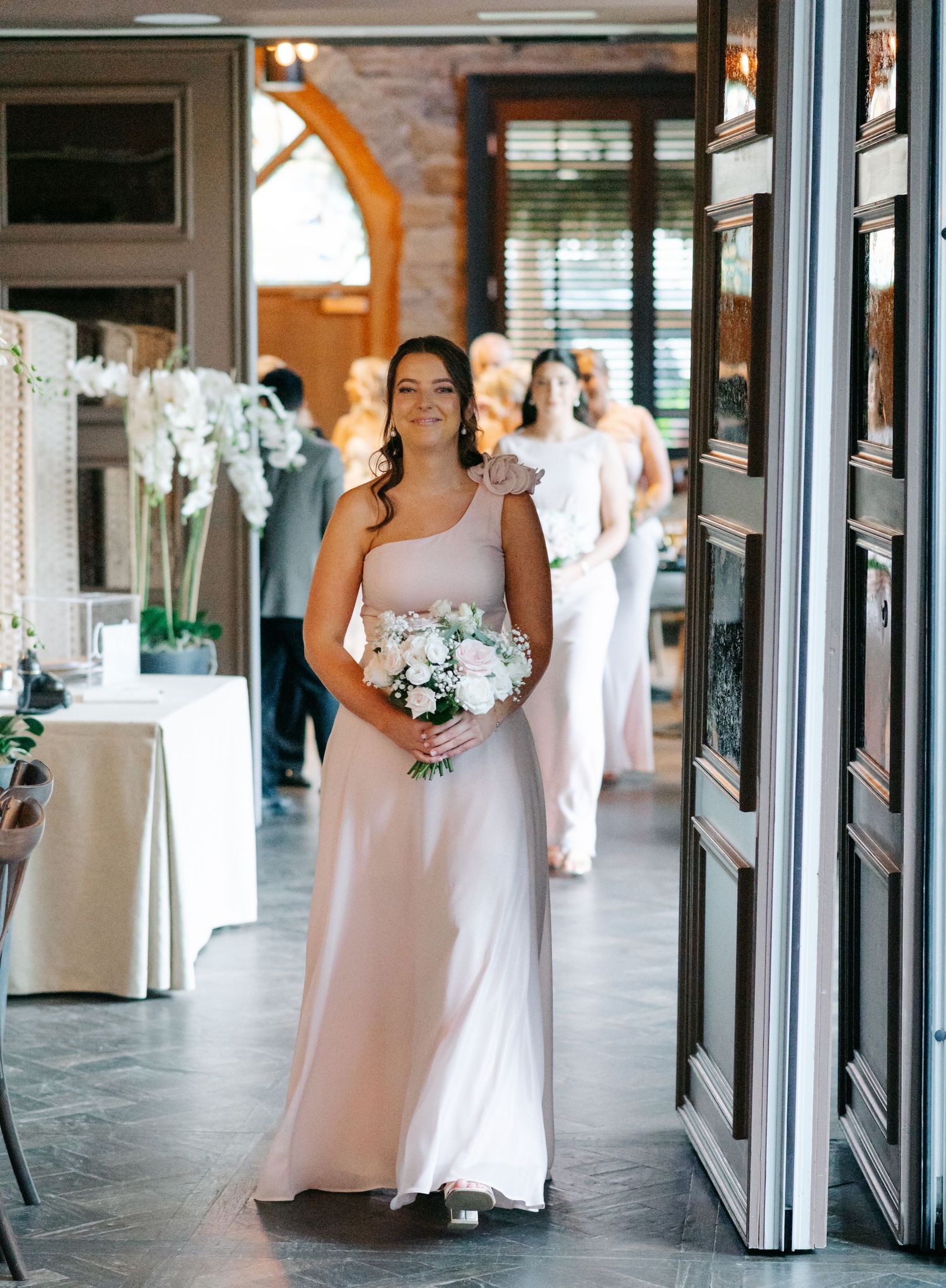 Bridesmaids in blush pink dresses walk through an elegant covered walkway holding white floral bouquets.