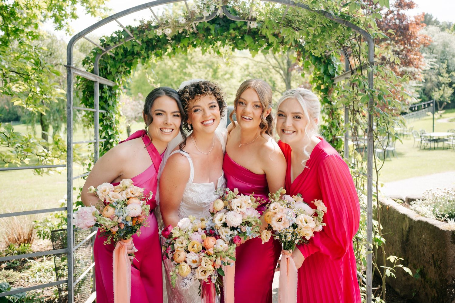 Bridesmaids in vibrant pink dresses pose with floral bouquets under a garden archway on a sunny day.