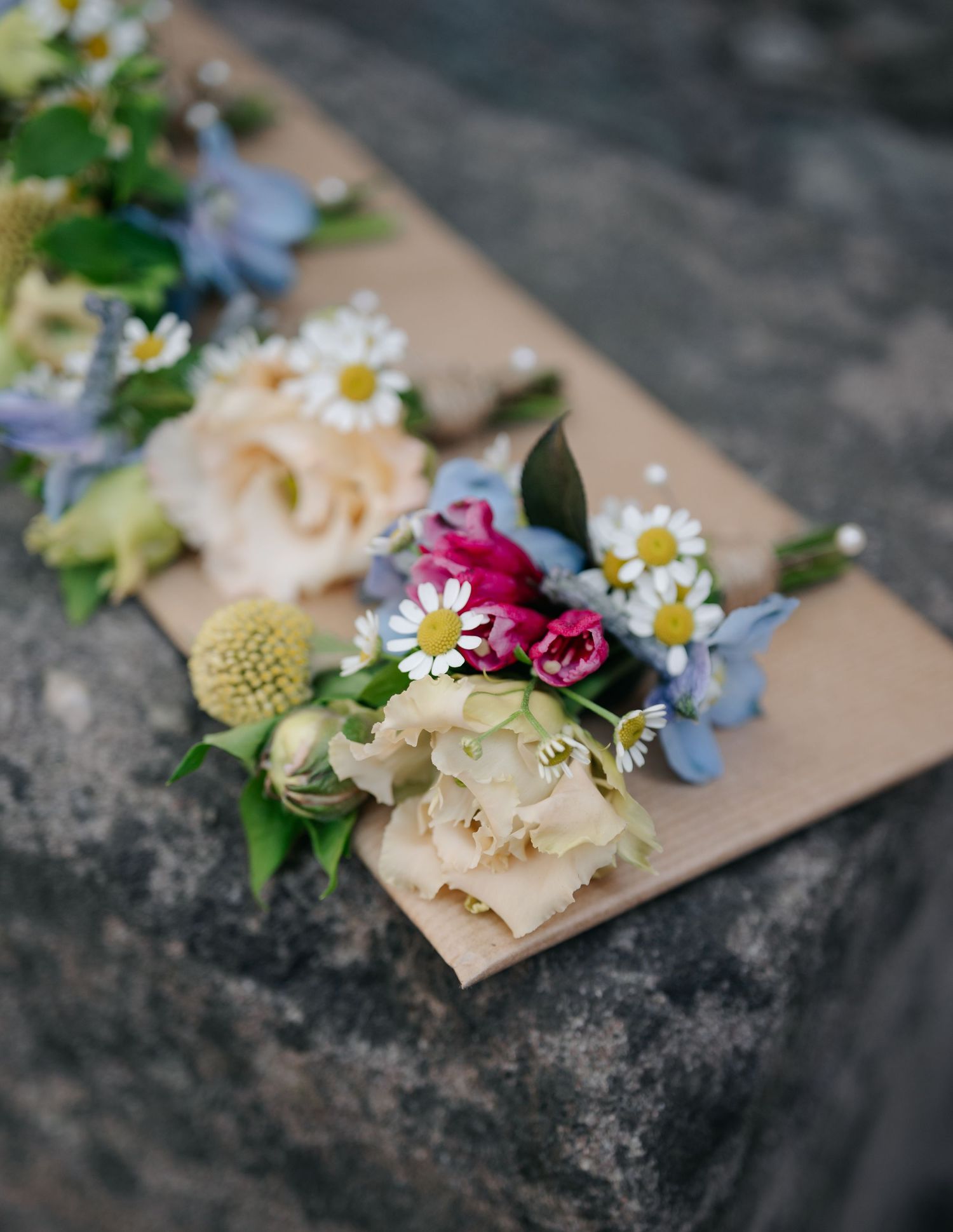Delicate floral arrangement with daisies, roses and blue flowers on a beige surface against stone background.