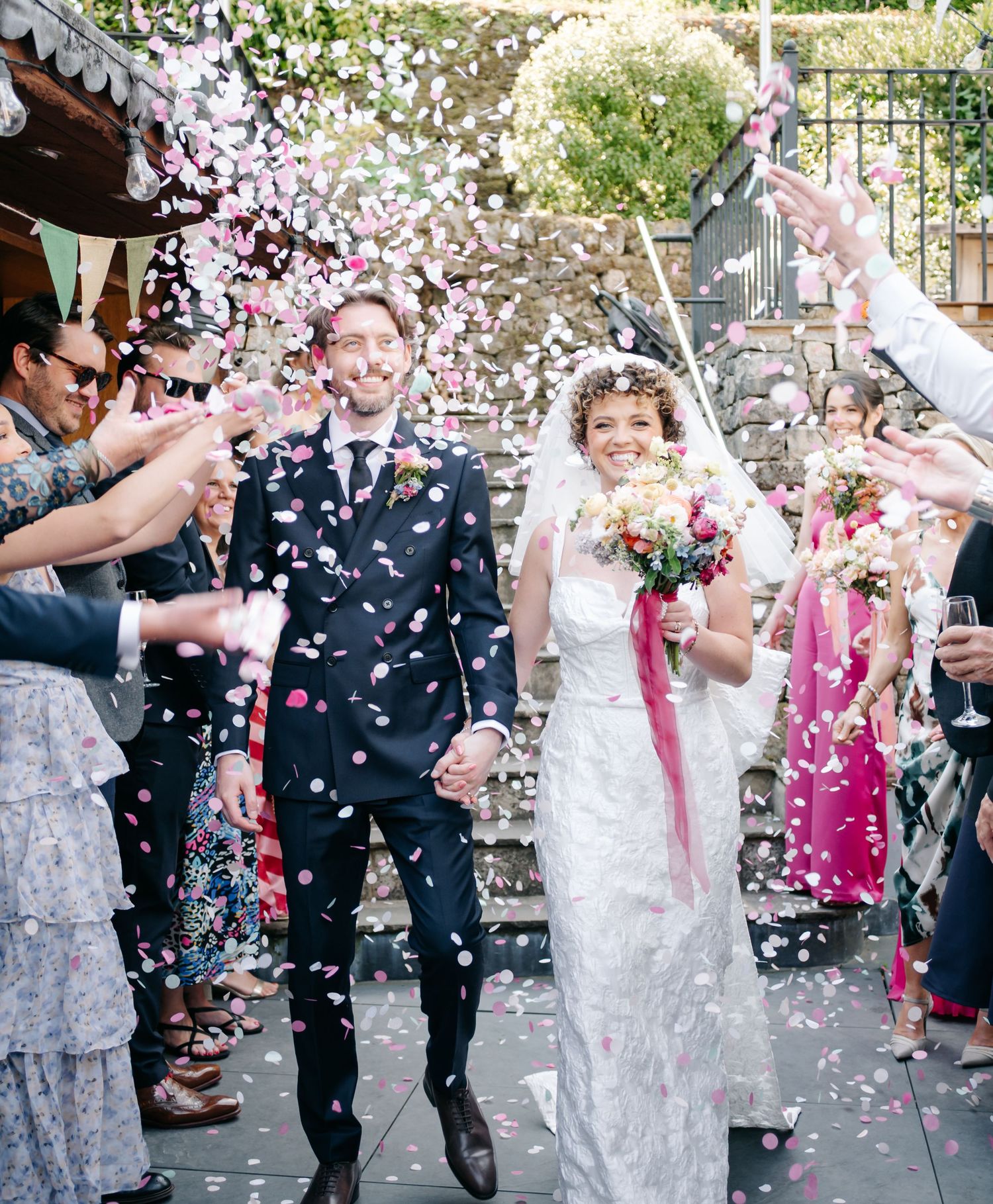Wedding party celebration with guests throwing white flower petals as confetti outdoors.