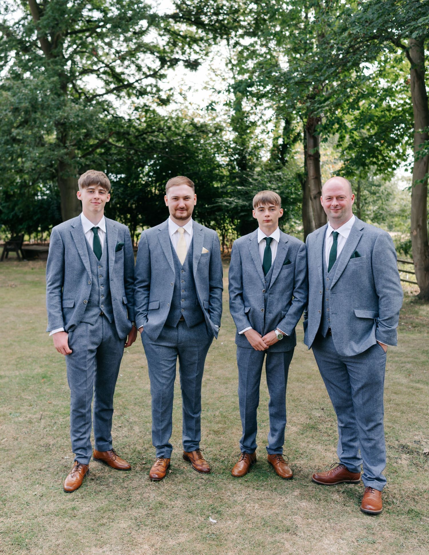 Four groomsmen in matching gray suits and brown shoes stand together on grass with trees in the background.