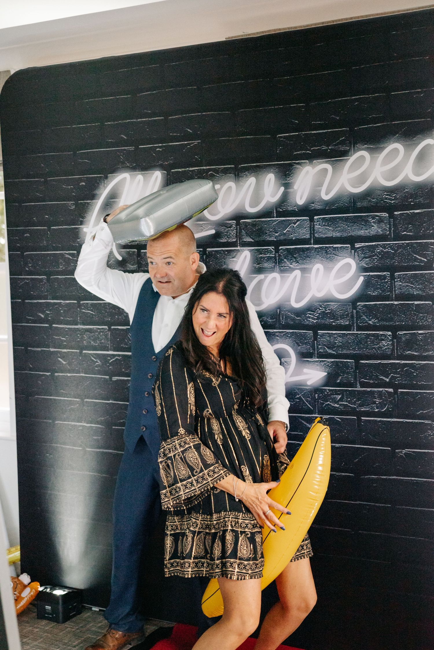Couple poses playfully against black brick wall with neon signage.