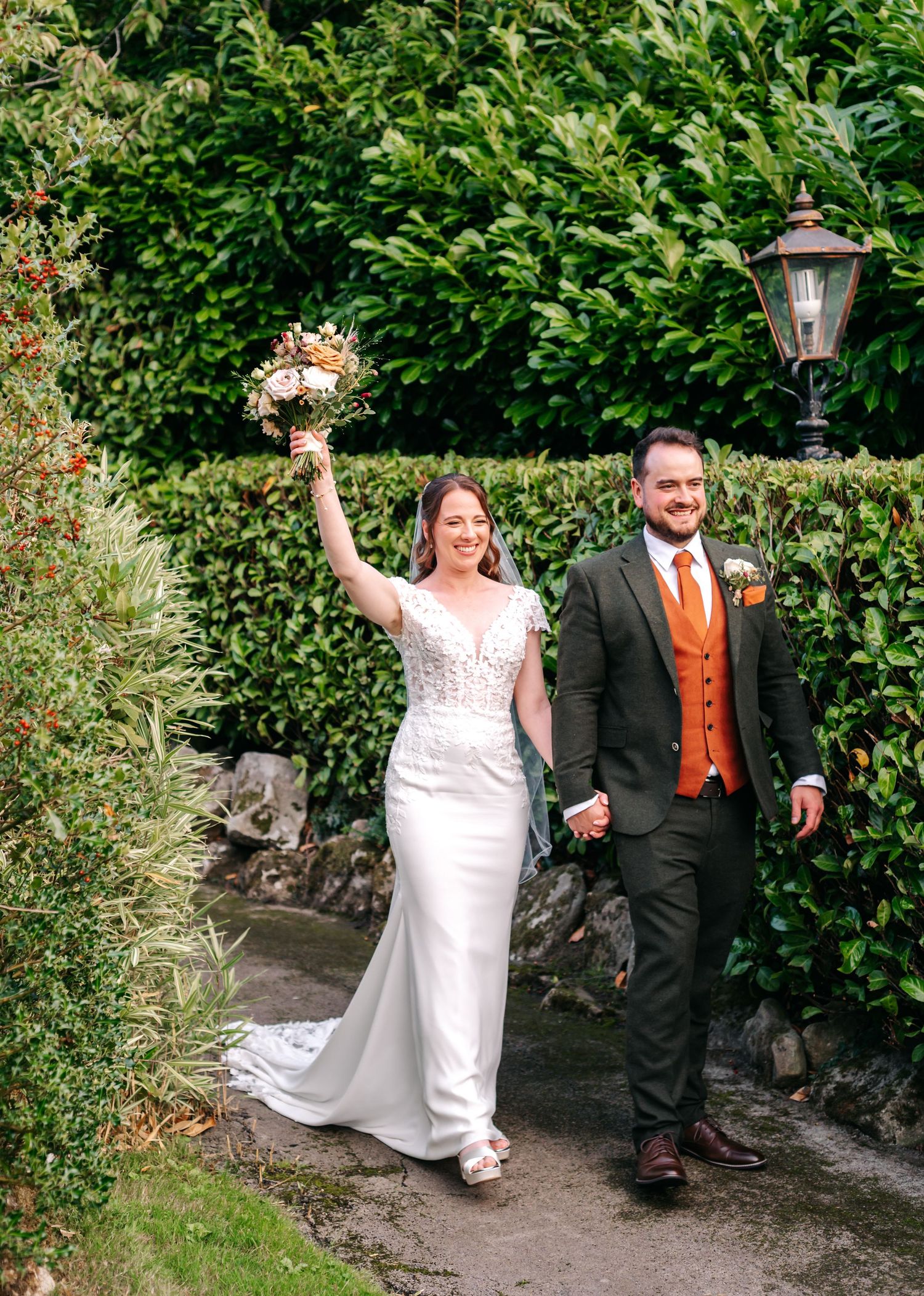 Smiling bride in a lace wedding gown raises her bouquet while walking beside the groom in an orange waistcoat along a garden path.
