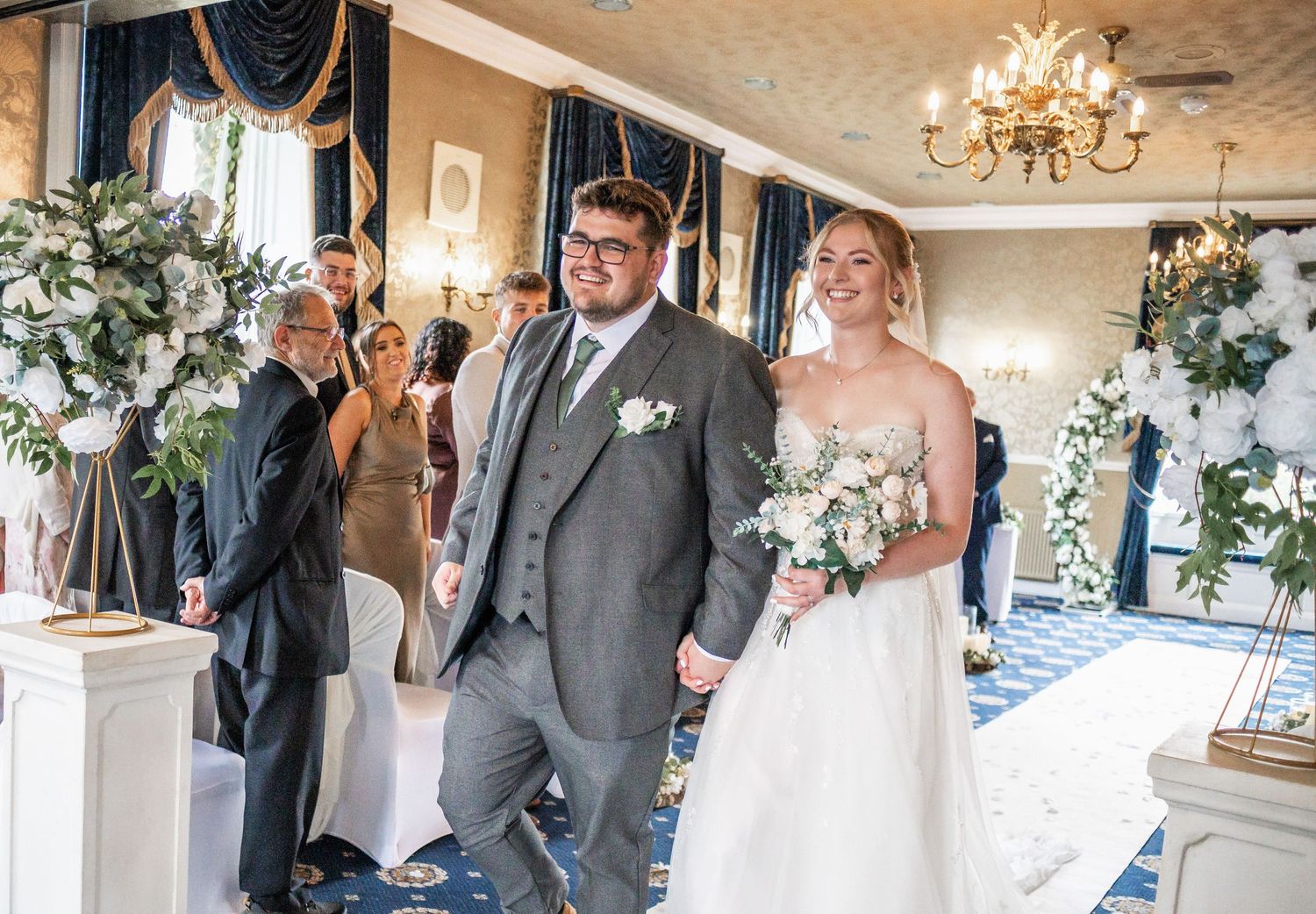 Wedding ceremony in an elegant room decorated with white floral arrangements and chandeliers.