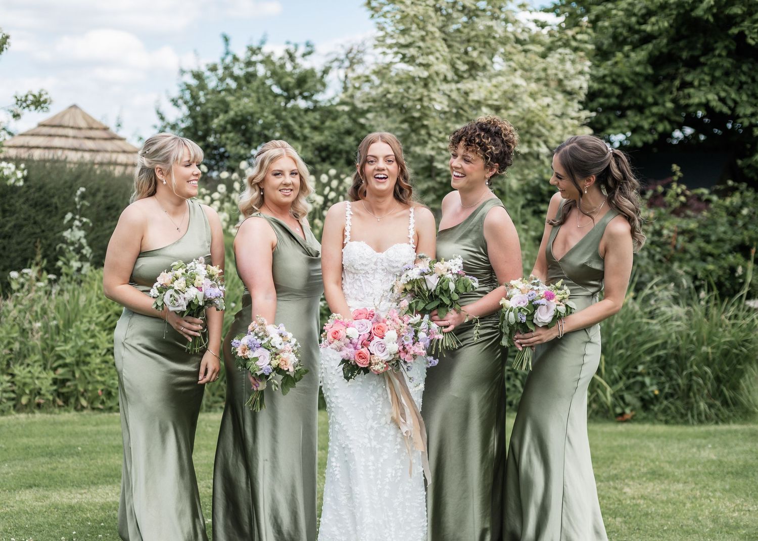 Wedding party wearing sage green dresses poses in elegant garden setting with summer flowers.